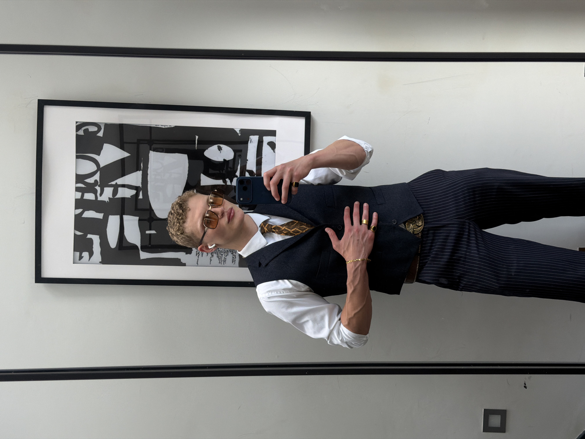 A young man with blonde curly hair is taking a mirror selfie in a room with white walls and a large framed abstract artwork on the wall. He is wearing a white dress shirt, a dark grey vest, a patterned tie, dark pinstripe pants, a gold belt with a large ornate buckle, and gold rings on his fingers. He also has on sunglasses and AirPods. He is holding a dark blue smartphone in his right hand, and his left hand is resting on his chest. The room appears to be a dressing room or a fitting room, possibly in a retail store in London, United Kingdom, given the style of the artwork and the modern aesthetic.