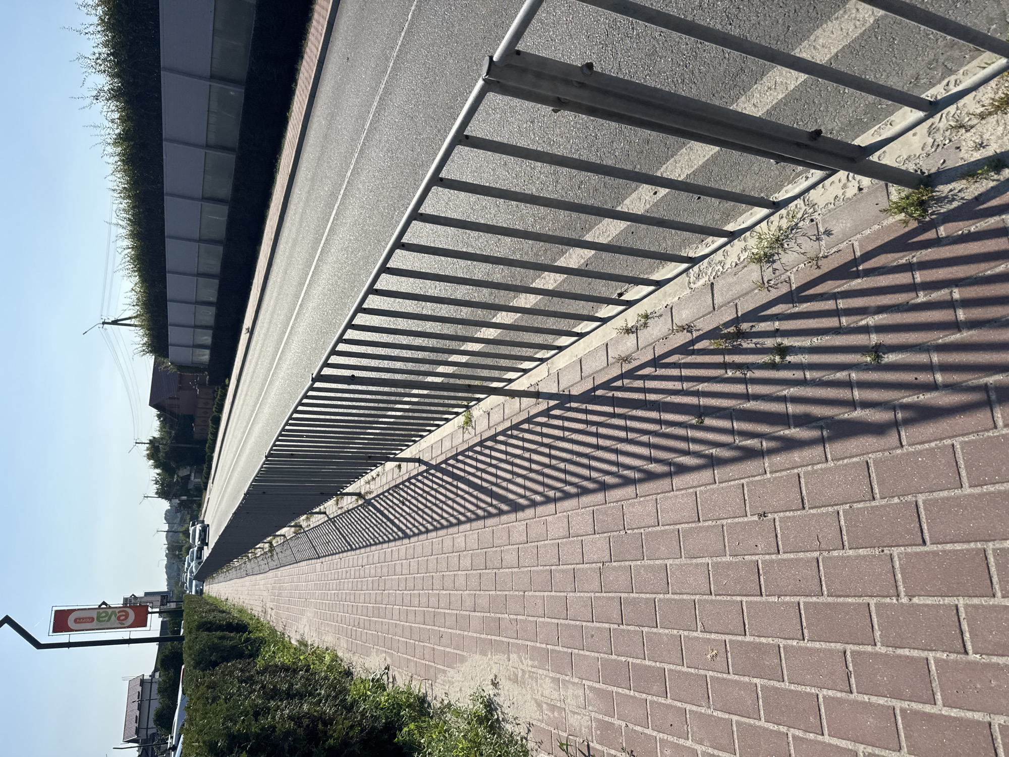 A sunlit outdoor scene on a clear day depicts a paved sidewalk running alongside a road. The sidewalk is made of red brick pavers, and a metal fence with vertical bars separates it from the road. The fence casts long, striped shadows on the sidewalk due to the low angle of the sun. Green hedges line the left side of the sidewalk, and on the right side, beyond the fence, the road is visible with two lanes and white markings. In the distance, there are buildings and trees, and a tall pole with a red banner hangs on the left. Several cars are visible on the road, suggesting traffic. The overall environment appears to be an urban or suburban roadside.