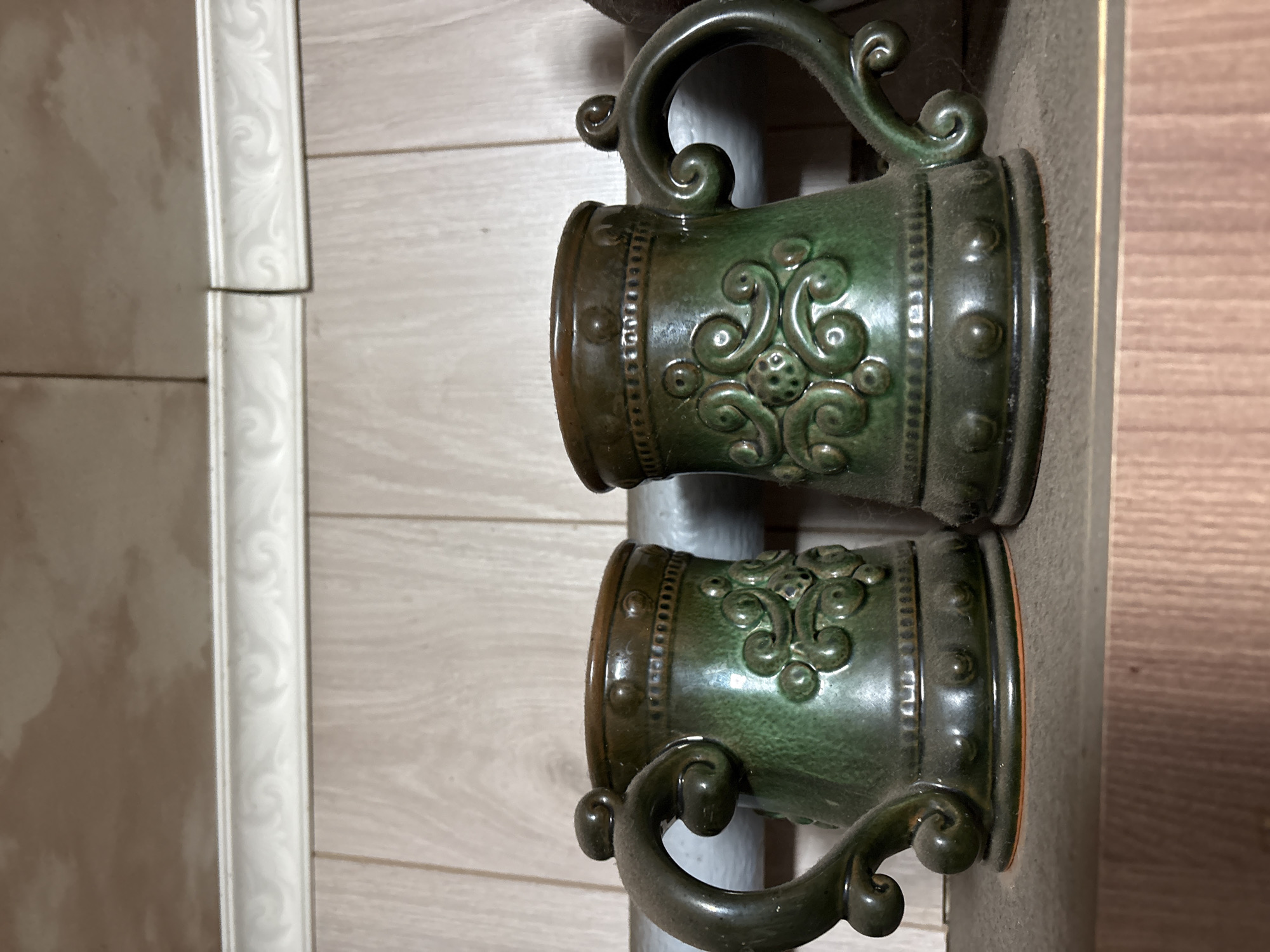 Two green ceramic mugs with ornate relief decorations and handles are displayed on a shelf. The mugs have a rustic appearance, with visible dust accumulation. The background consists of wooden paneling and a decorative white trim along the top of the wall. The scene suggests an indoor setting, possibly a home or a shop, in the city of Vetluzhsky, Russia.