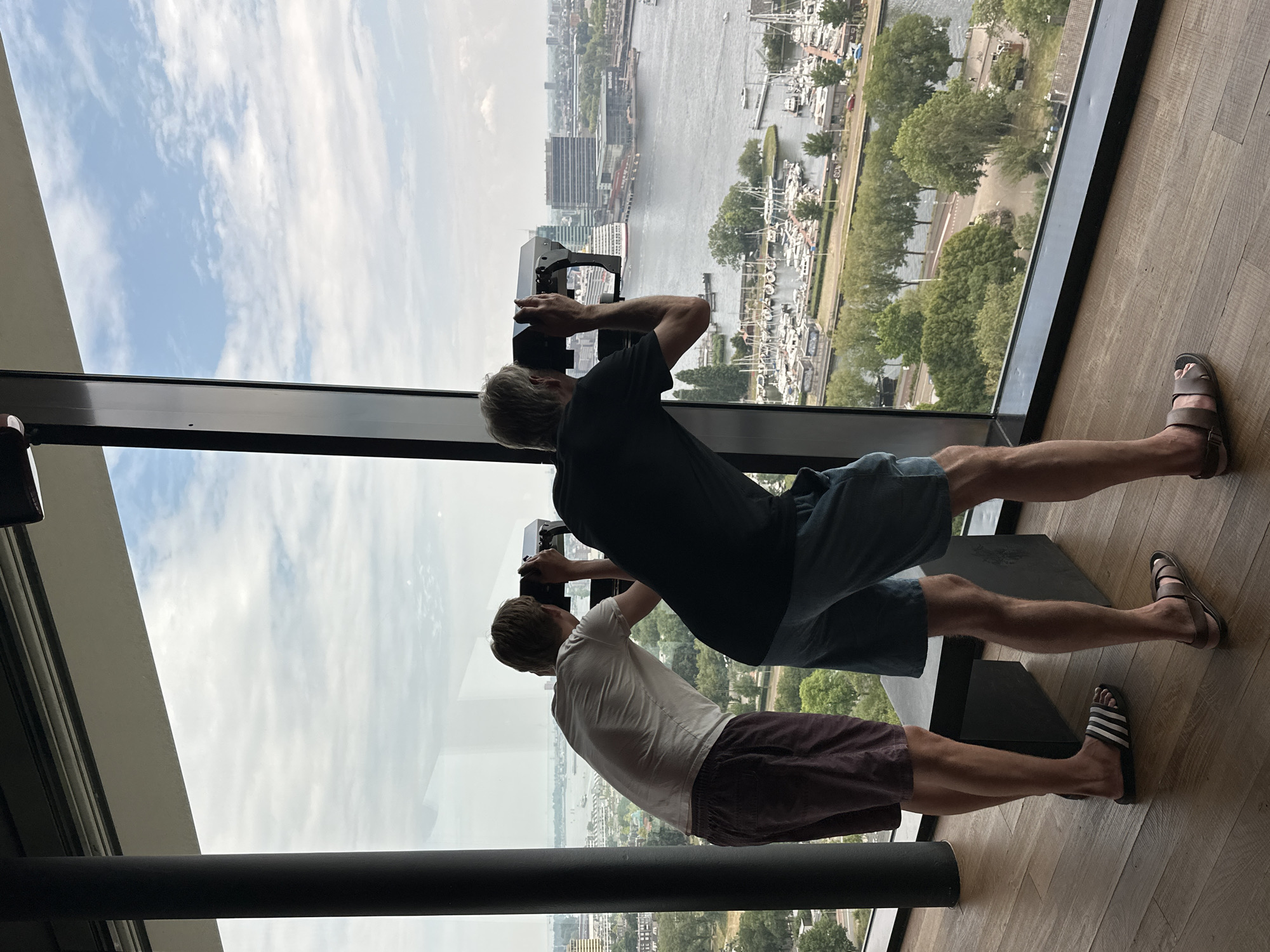 Two individuals are standing indoors, looking out through a large window at a cityscape. The individual on the left, wearing a white t-shirt and burgundy shorts, is facing away from the camera and appears to be looking through a viewing scope. The individual on the right, wearing a dark t-shirt and blue shorts, is also facing away and is actively using a viewing scope. The window offers a panoramic view of a harbor filled with numerous boats, a body of water, and buildings in the distance. Trees and greenery are visible between the viewer and the harbor. The floor inside is made of polished wood. The setting appears to be a high vantage point in Amsterdam, Netherlands, likely an observation deck or a building with a scenic view.