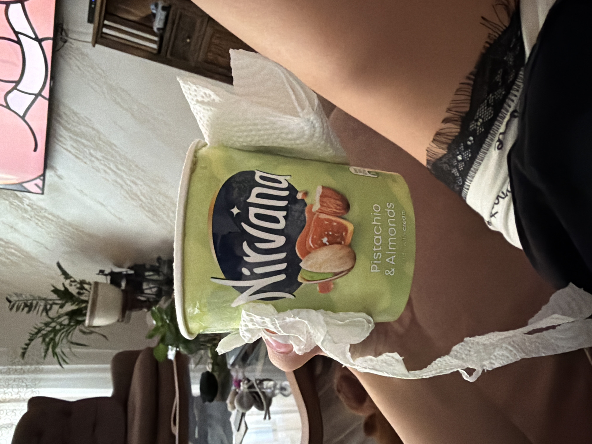 A person is holding a container of Nirvana Pistachio & Almonds ice cream. The container is wrapped with paper towels. The person is wearing a white shirt with black lace trim and black shorts. The background shows a partially visible TV screen, a wooden cabinet, plants, and a table with decorative items. The location is not identifiable as Gabrovo, Bulgaria, from the visual data.