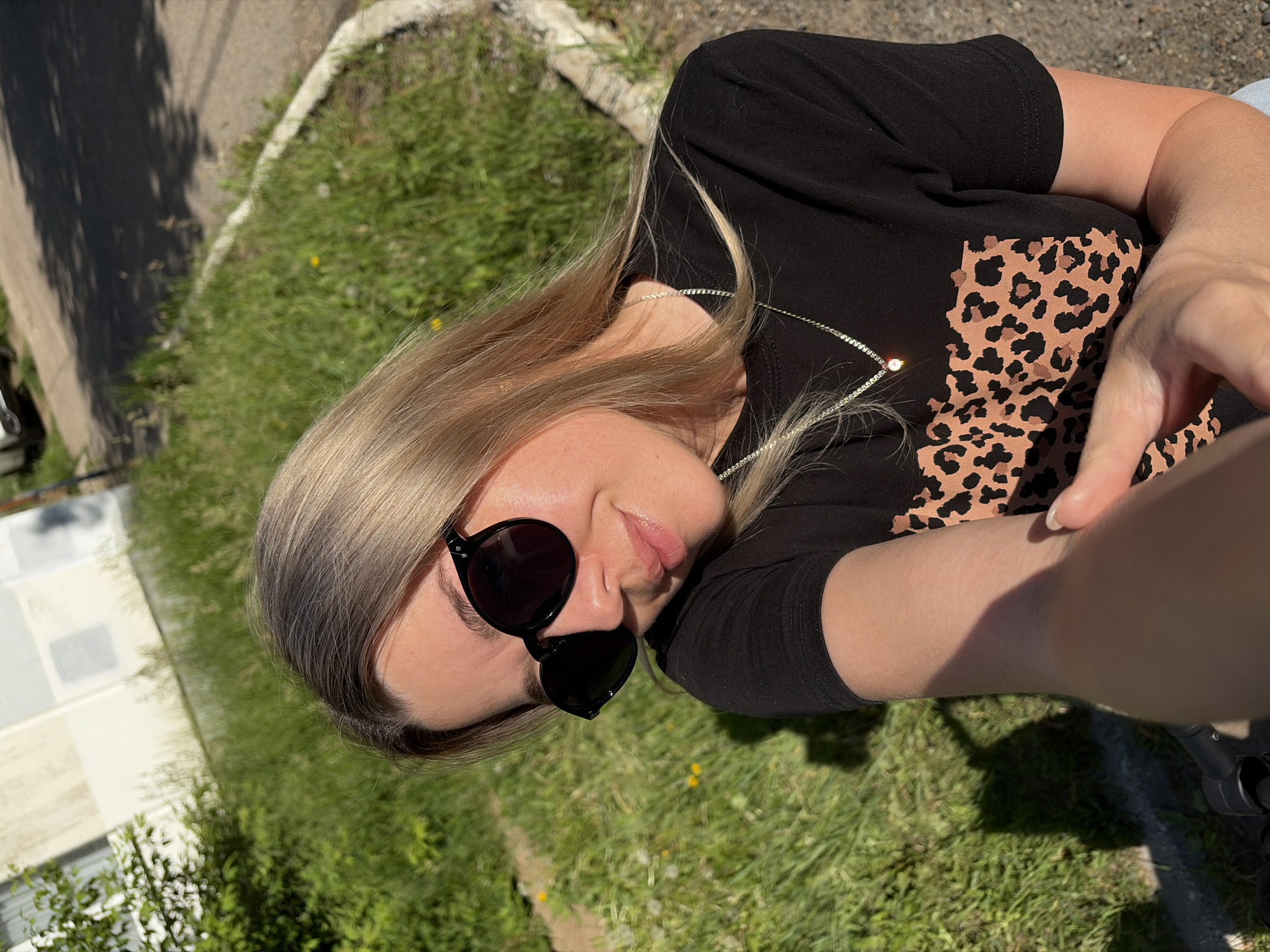 A blonde woman with sunglasses is taking a selfie outdoors on a sunny day. She is wearing a black t-shirt with a leopard print design and a silver chain with a diamond pendant. The background shows a grassy area next to a paved path, with a building visible in the distance. The scene appears to be in the city of Chaikovsky, Russia, as suggested by the context. The woman is smiling and looking at the camera.