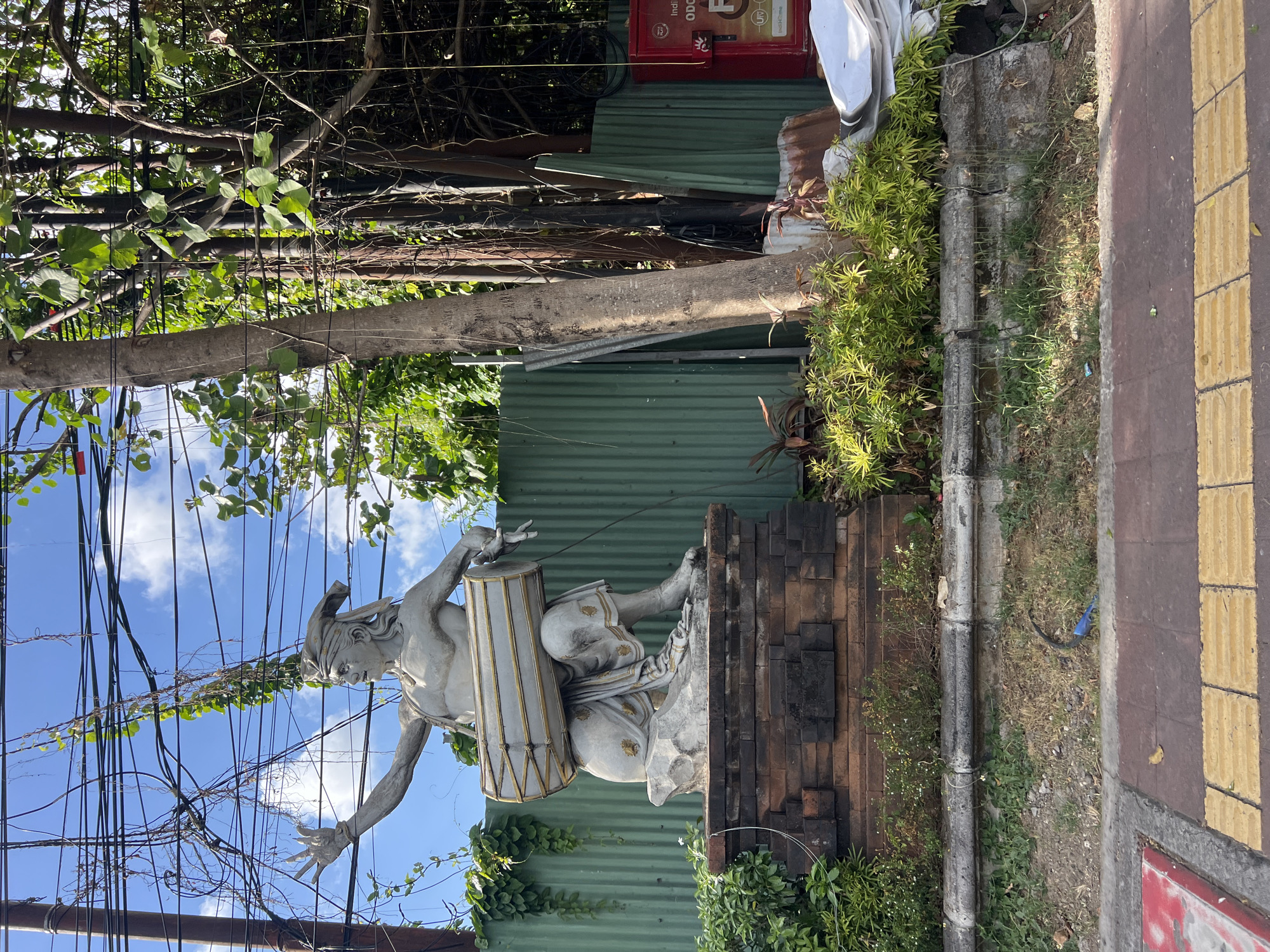 A statue of a Balinese musician playing a drum is situated in front of a corrugated metal fence in Denpasar, Indonesia. The statue is made of concrete and depicts a man seated with a drum across his lap, holding drumsticks. He wears traditional Balinese clothing and a headscarf. The statue is mounted on a brick pedestal, surrounded by greenery. Overhead, a tangle of electrical wires crisscrosses the sky, adding a layer of urban clutter to the scene. In the background, a red utility box with markings is visible. The area appears to be a public space, with a sidewalk in the foreground featuring tactile paving for the visually impaired.