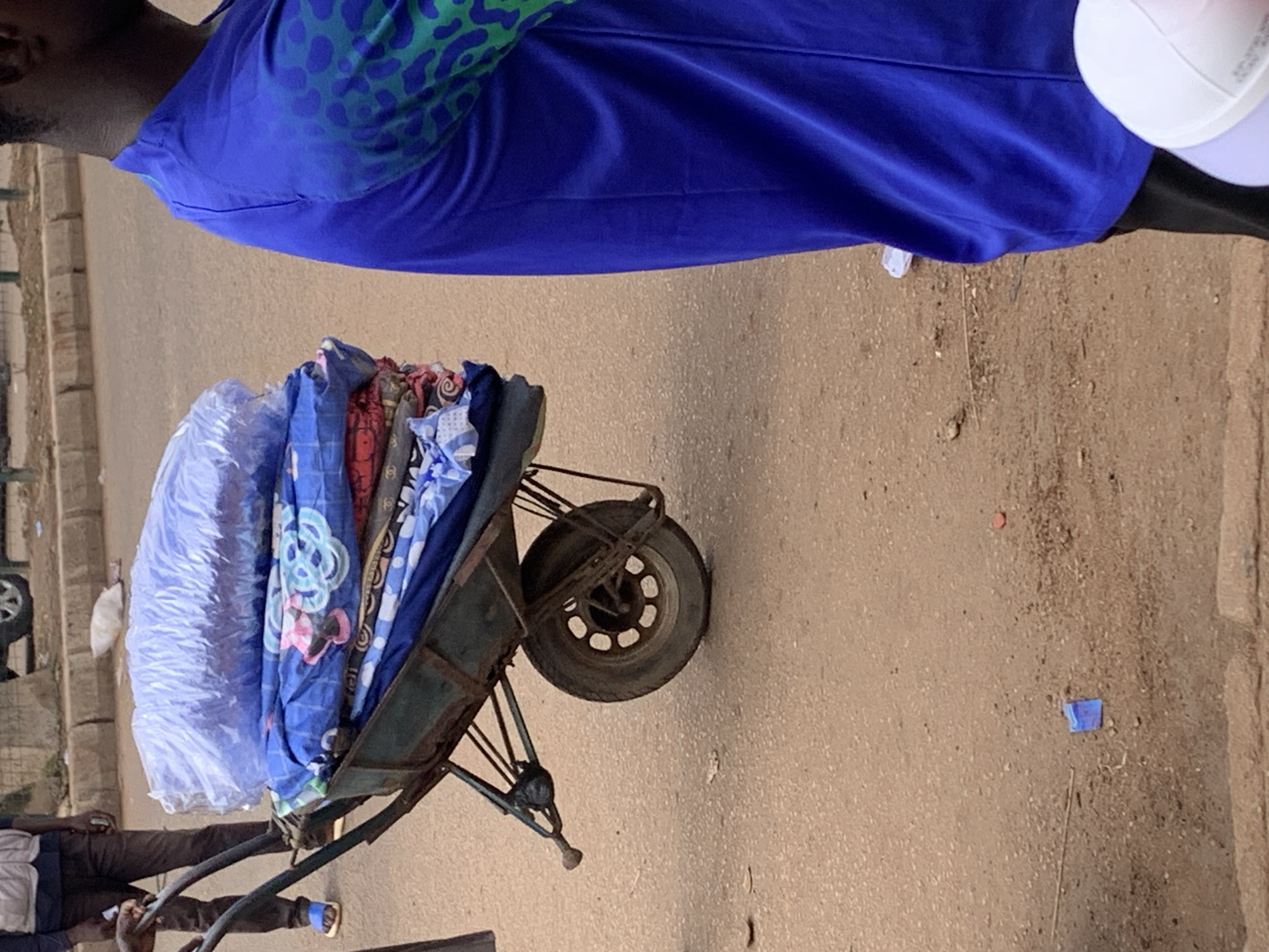 A person, likely a man, is shown from the waist up wearing a blue shirt with a green leopard-like pattern. He is standing next to a wheelbarrow filled with what appear to be textiles. The wheelbarrow is dark green and has a single wheel. The textiles are wrapped in plastic and several are visible with colorful patterns, including one with blue and white floral designs. The scene appears to be outdoors on a paved surface, possibly a street or market. The ground is a mix of dirt and asphalt. In the background, there are some brick structures and a parked car, suggesting an urban or semi-urban environment. A white cap or hat is visible on the right side of the image. The lighting suggests it is daytime. The overall impression is of a vendor or someone transporting goods.