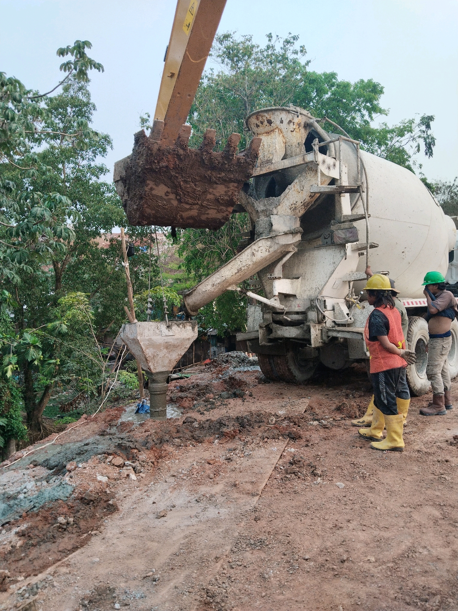 A concrete mixer truck is actively pouring concrete into a temporary chute that leads down to a construction site. Three construction workers are present. One worker, wearing a yellow hard hat, orange vest, and yellow boots, stands near the back of the truck. Another worker, wearing a green hard hat, stands behind the first worker. A third worker, partially obscured, is also near the truck. The environment is a construction area with loose soil, mud, and some vegetation. The scene suggests ongoing construction work, likely for a foundation or support structure, given the way the concrete is being directed.