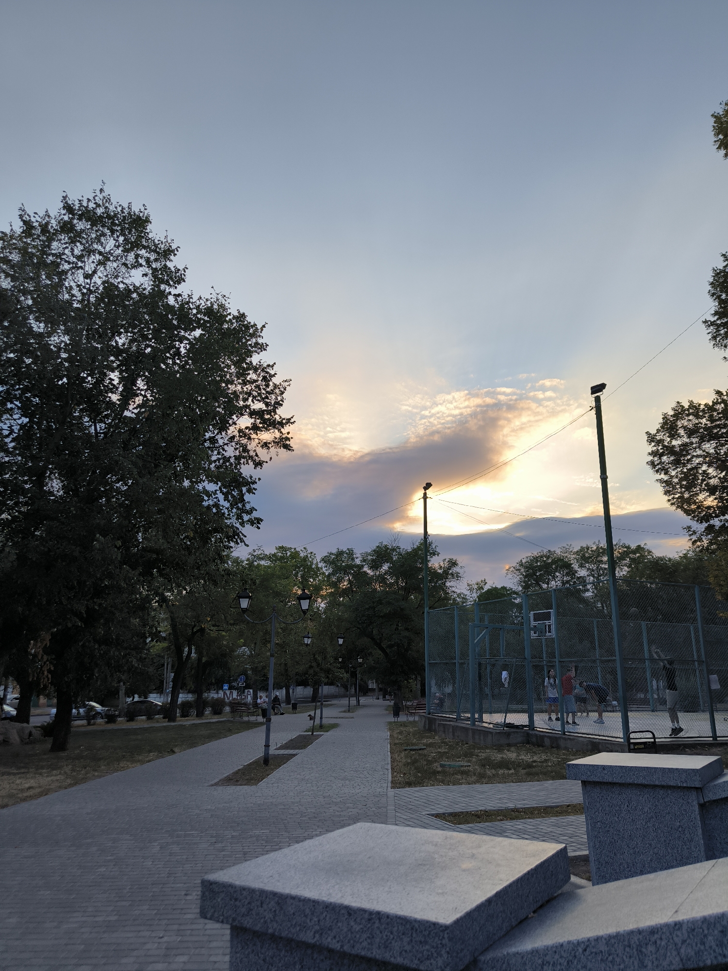 The image shows a park scene at dusk in Mykolaiv, Ukraine. A paved walkway runs through the park, lined with trees and streetlights. In the background, a basketball court with a fence is visible, and several people are playing basketball. The sky is a pale blue with some clouds catching the last rays of the setting sun. The overall atmosphere is peaceful and recreational.