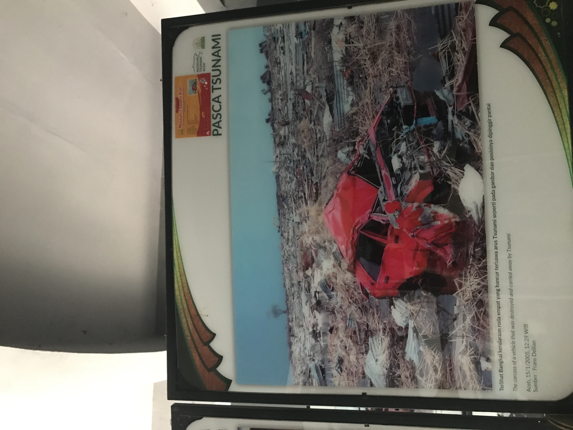 The image shows a framed photograph depicting the aftermath of the 2004 tsunami in Banda Aceh, Indonesia. The photograph is titled "PASCA TSUNAMI," and it is displayed at the Museum Tsunami Aceh. The photograph shows a red, four-door vehicle that is destroyed and carried away by the tsunami. The car is surrounded by debris. The photo is labeled: "The carcass of a vehicle that was destroyed and carried away by Tsunami Aceh, 15/1/2005, 12:29 WIB Sumber: Frans Dellian".