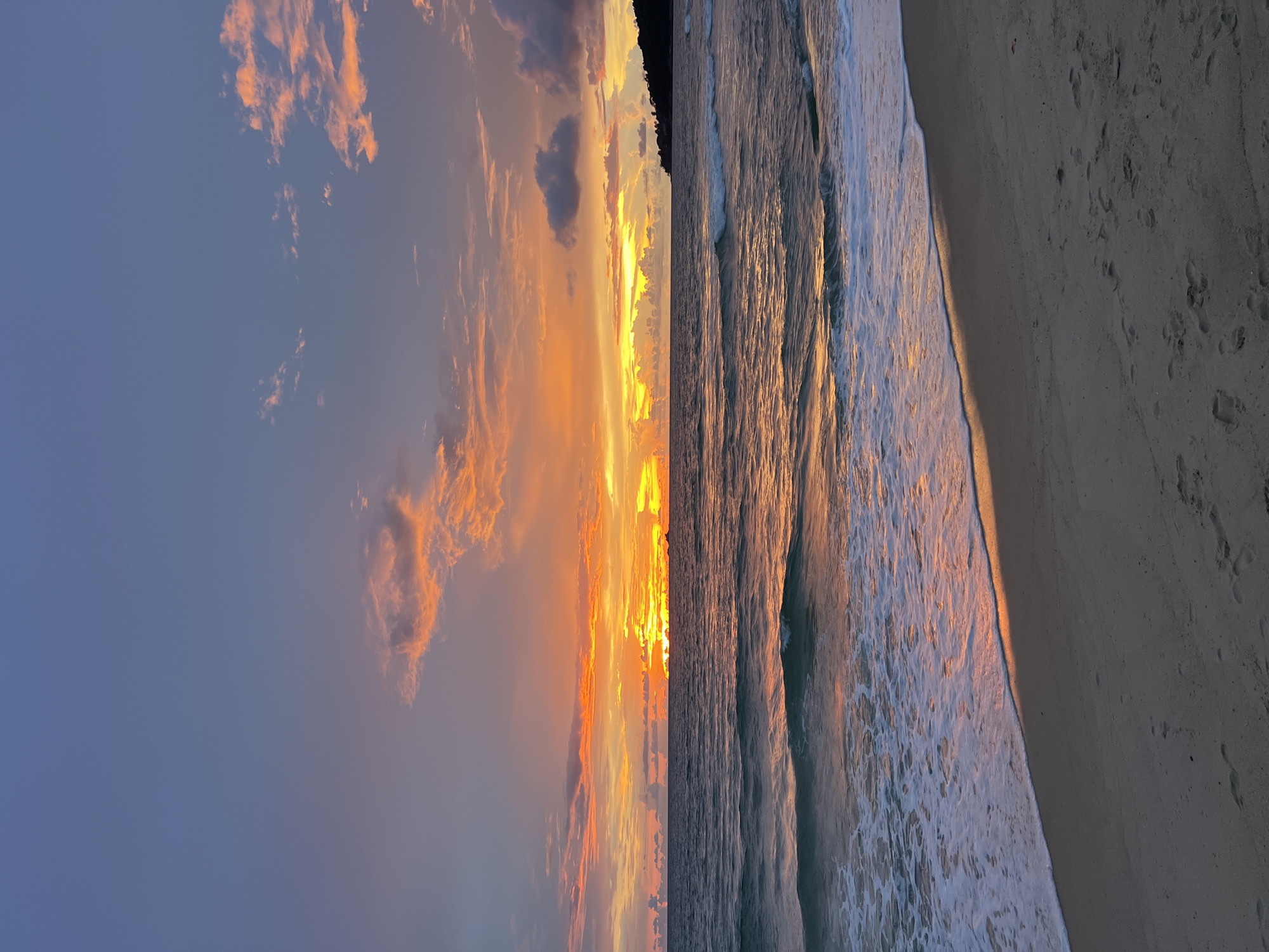 The image shows a beach scene at sunset. The sky is filled with vibrant colors of orange, yellow, and purple, with scattered clouds. The sun is setting over the ocean, casting a warm glow on the water. Gentle waves are rolling onto the sandy shore. In the distance, a silhouetted landmass can be seen along the horizon. The sand on the beach has footprints, indicating recent human presence. The overall scene is serene and picturesque.