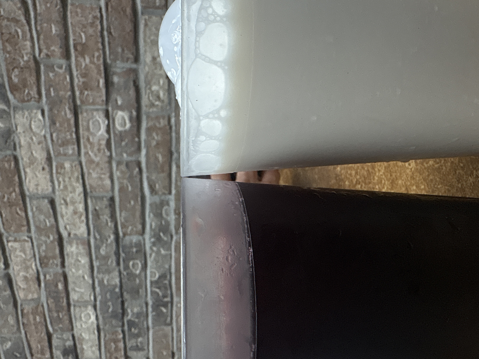 The image shows two cylindrical containers, one white and one dark, positioned adjacent to a brick wall. The white container appears to be filled with a frothy liquid. The dark container is filled with a darker substance and exhibits condensation. The brick wall is weathered and composed of rectangular bricks. The location is unknown and cannot be identified from the image.