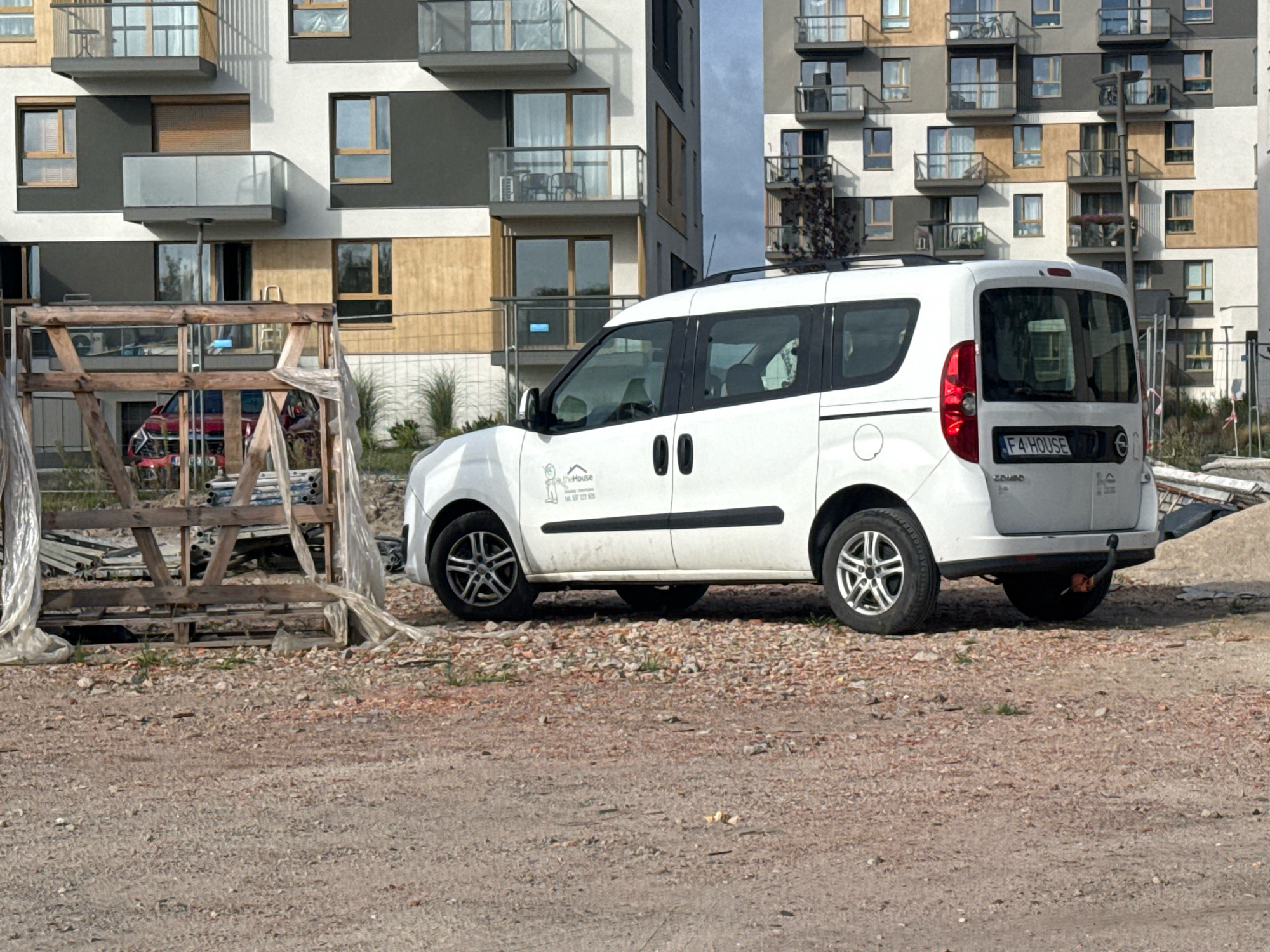 A white minivan with "F4 HOUSE" license plate is parked on a gravel lot in front of a modern apartment building in Nowa Sól, Poland. The building has a contemporary design with balconies and large windows. To the left of the van, there is a wooden structure covered with plastic sheeting, and a red car is partially visible behind it. The ground is covered with gravel and some loose stones. The sky is overcast.