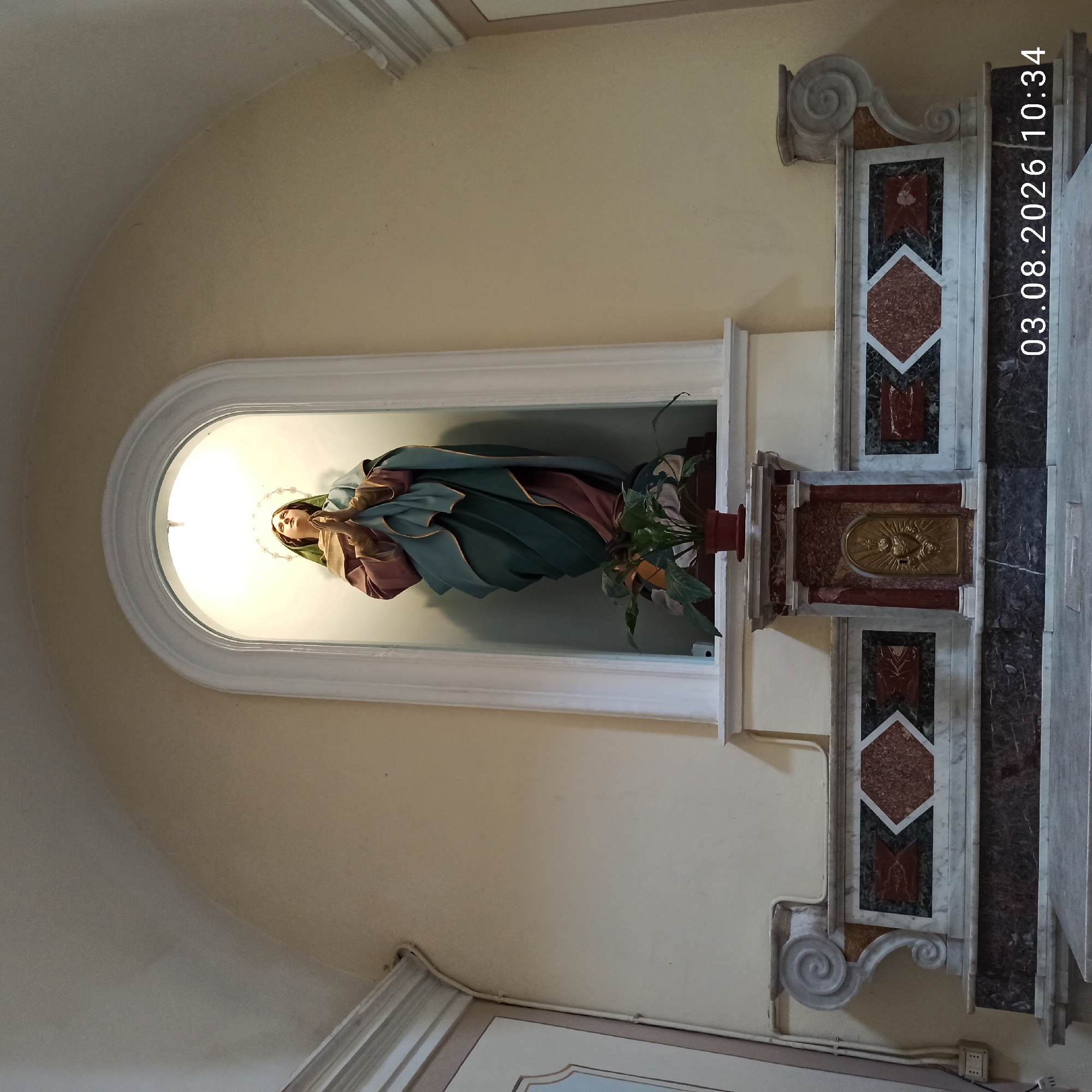 The image shows a statue of a praying woman, likely the Virgin Mary, in an arched niche above an altar. The statue is dressed in flowing robes of blue-green and mauve, with a green head covering. A halo is visible behind her head, and her hands are clasped in prayer. A potted plant sits at the base of the statue.
The altar is made of marble and features ornate decorative elements, including geometric inlays of various colored stones and a central gold relief of a heart with a cross. The walls are a pale yellow color, and the ceiling is arched.
The lower right corner of the image displays a timestamp: "03.08.2026 10:34".
This statue and altar are located in the church of San Rocco in Ruvo di Puglia, Italy. The church, built in the late 16th century, houses several important works of art.