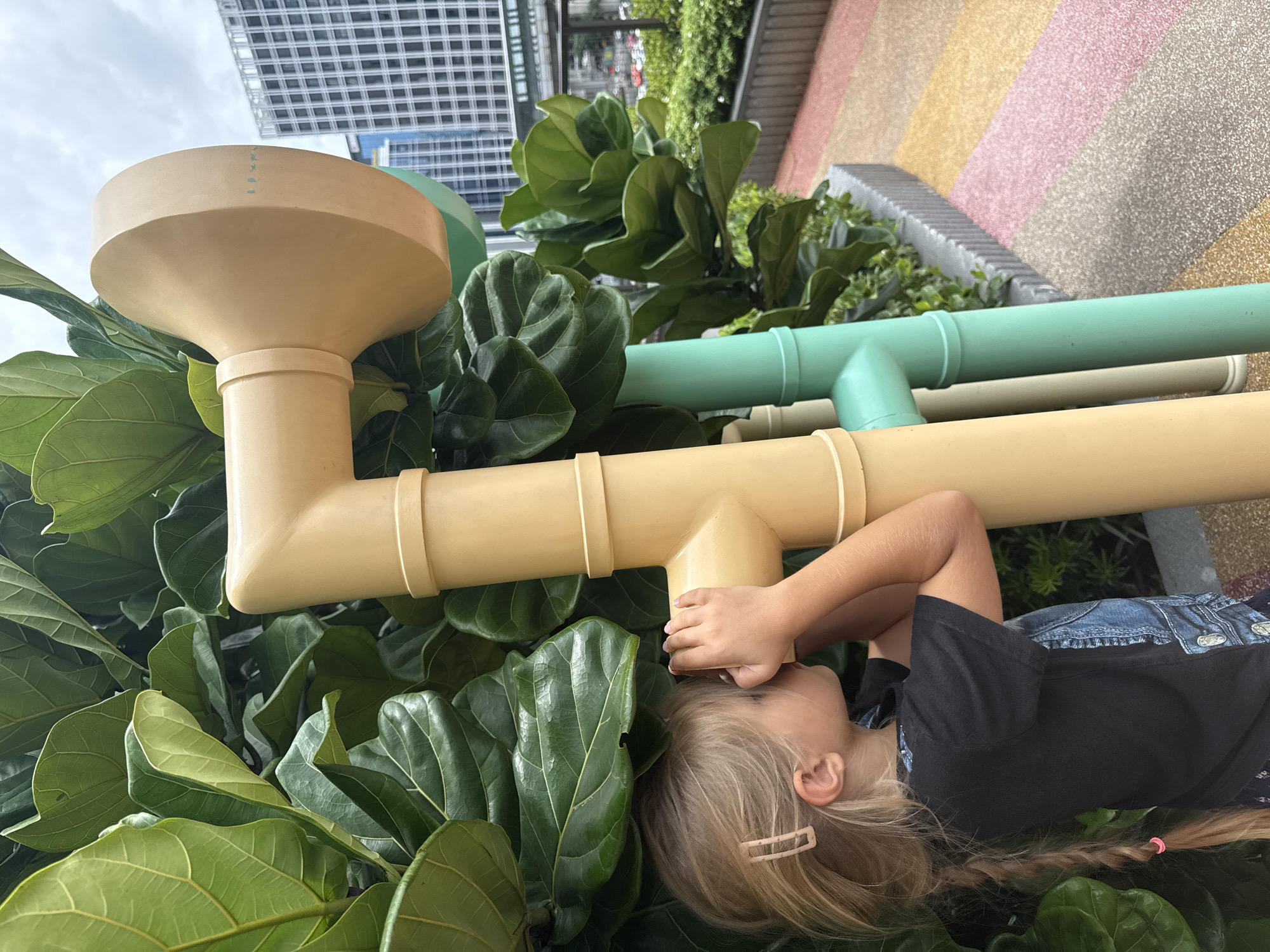 A young girl with blonde hair in a braid, wearing a black t-shirt, looks through a large beige pipe-like structure. She is standing outdoors, with large green leaves of a plant to her left. In the background, there are tall buildings and a cloudy sky, suggesting an urban environment. The ground is a colorful patterned walkway. This scene appears to be in a public space within Bangkok, Thailand.