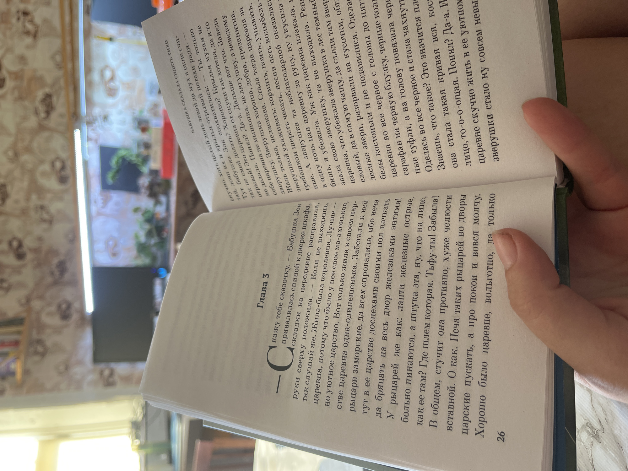 The image shows a close-up of an open book, held by a person's hand. The book is in Russian, with text on both pages. The right page has a title at the top: "БАБУШКА СКАЗАЛА СИДЕТЬ ТИХО" (Grandmother said to sit quietly). The left page has a chapter heading: "Глава 3" (Chapter 3) and a large, stylized letter "C" at the beginning of a paragraph. The book is lying flat, with the spine slightly open. The background is blurry but appears to be an indoor setting with wallpaper and some electronic devices. The lighting suggests it might be daytime, possibly near a window. There are no discernible landmarks or indications of Krasnyye Barrikady, Russia, in the image itself.