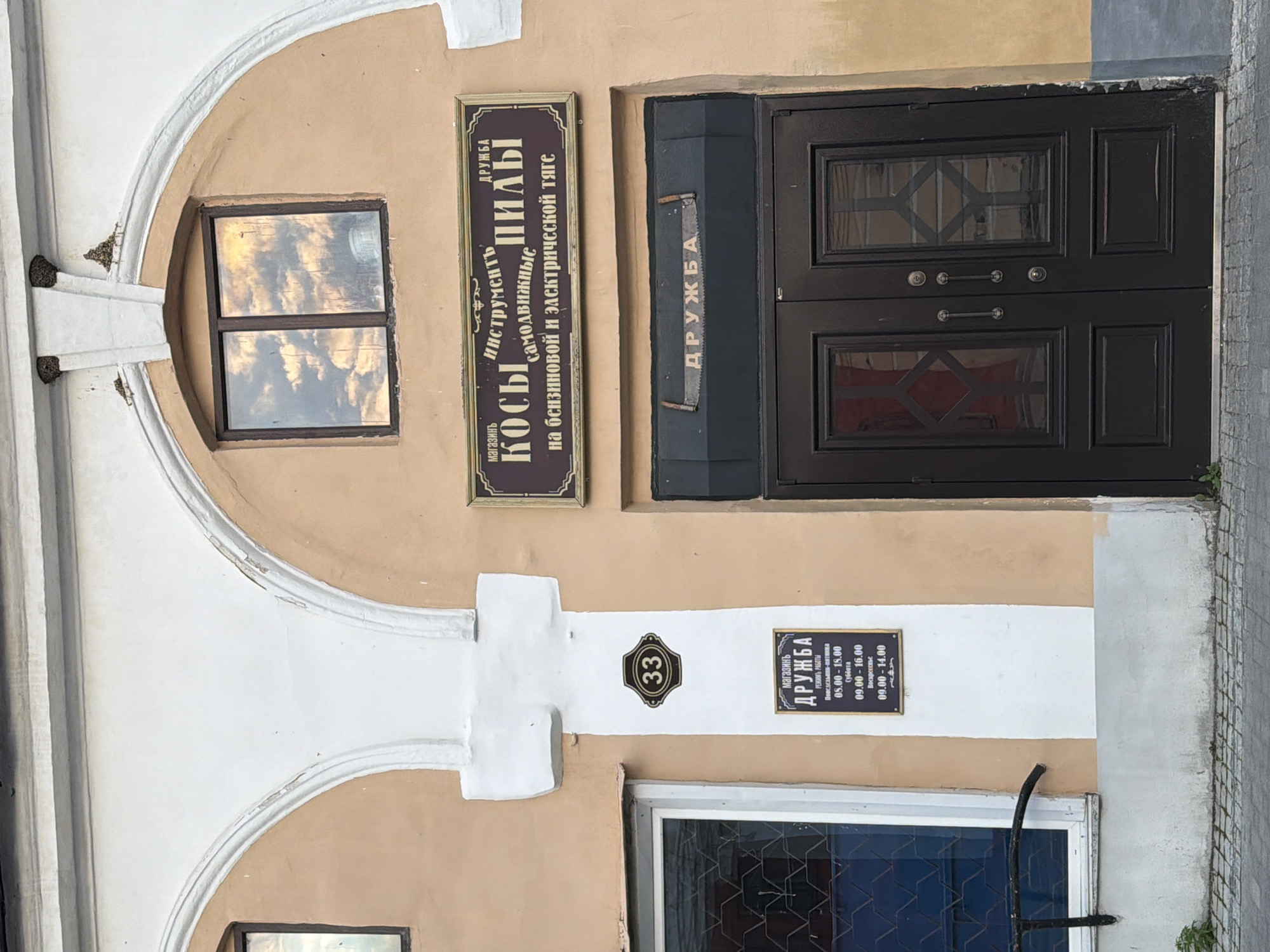 The image shows the facade of a shop called "Drugba" in Arzamas, Russia. The shop is located in a building with a light-colored archway and beige walls. A sign above the entrance reads "магазин ИНСТРУМЕНТс ДРУЖБА КОСЫ пилы самодвижные на бензиновой и электрической тяге," which translates to "Shop Tools DRUZHBA MOWERS SAWS self-propelled on gasoline and electric traction." There is a smaller sign with "33" on it. Below the main sign, a saw with the word "ДРУЖБА" written on it is displayed above a set of double doors. A sign to the left of the entrance lists the shop's operating hours. The image is taken from a low angle, with the sky reflected in a window above the entrance.