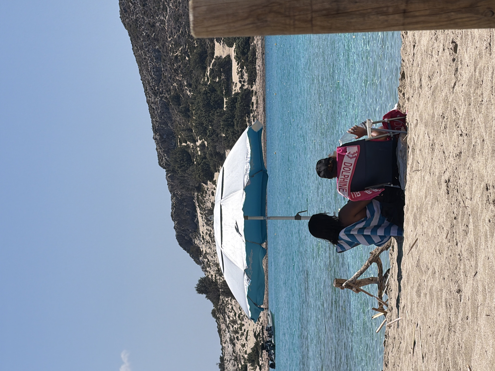 In an outdoor scene on a sandy beach in Greece, two individuals are seated under a large, open umbrella. The person on the left, facing away from the camera, is wrapped in a blue and white striped towel. The person on the right, also facing away, is wearing a dark cap and a pink and black shirt with the word "DOLPHINE" printed on the back. They are holding a clear bottle in their right hand. The umbrella is positioned over them, providing shade from the sun. In the background, the turquoise sea is visible, with a rocky, sparsely vegetated hillside rising behind it. A few boats are moored in the water on the left side of the frame. A wooden post stands in the foreground on the right, partially obscuring the view. The overall setting suggests a tranquil day at the beach.