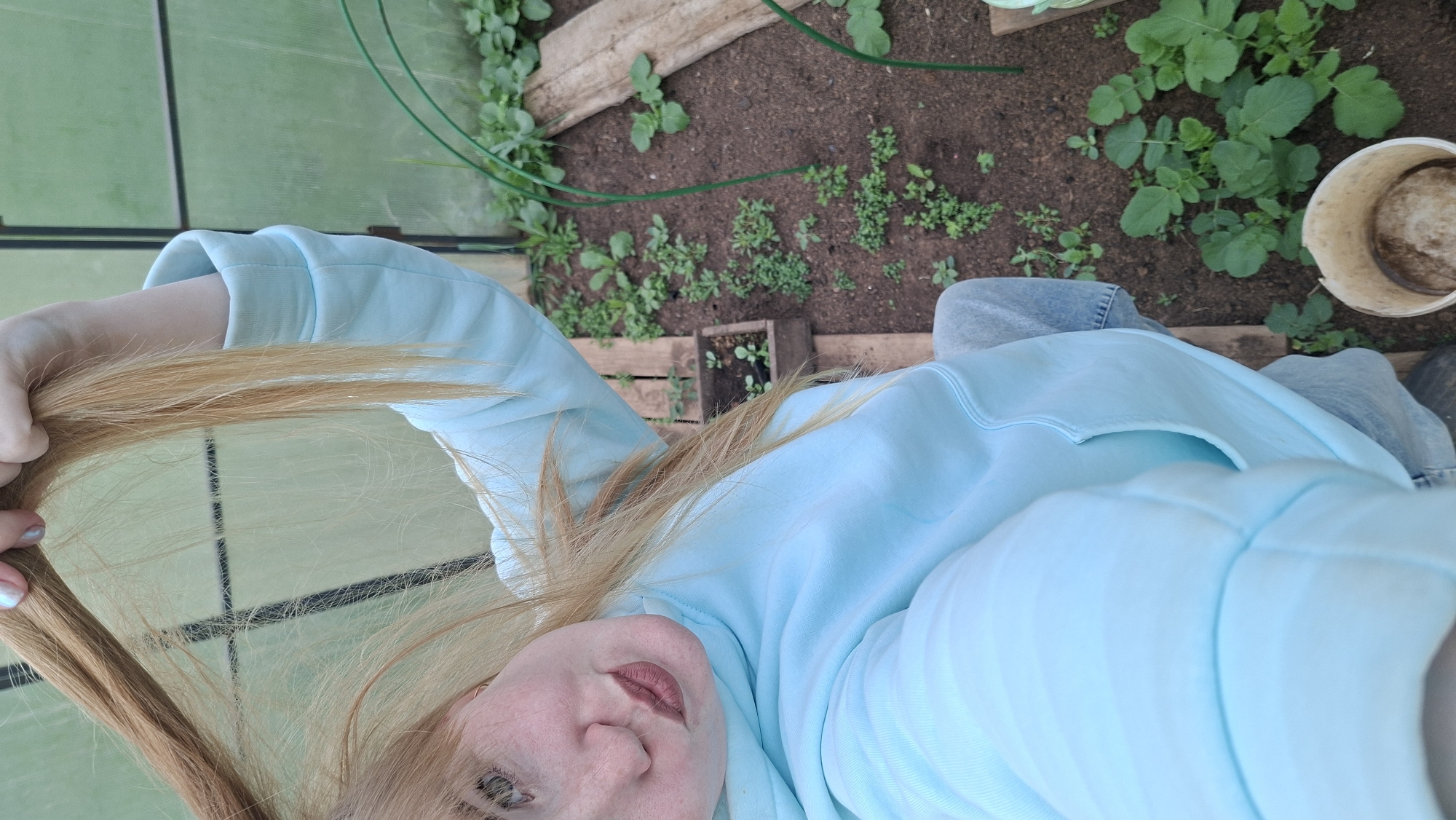 A selfie was taken inside what appears to be a greenhouse or plant growing structure. The photographer, a fair-skinned person with red hair, is wearing a light blue sweatshirt and jeans. The selfie captures the top portion of their face and the hair they are holding.
The environment shows a planted area with various plants and seedlings. The soil is dark. A white bucket or container is visible on the right side of the frame. The greenhouse structure has transparent or semi-transparent material forming the walls and roof, with black structural beams visible.