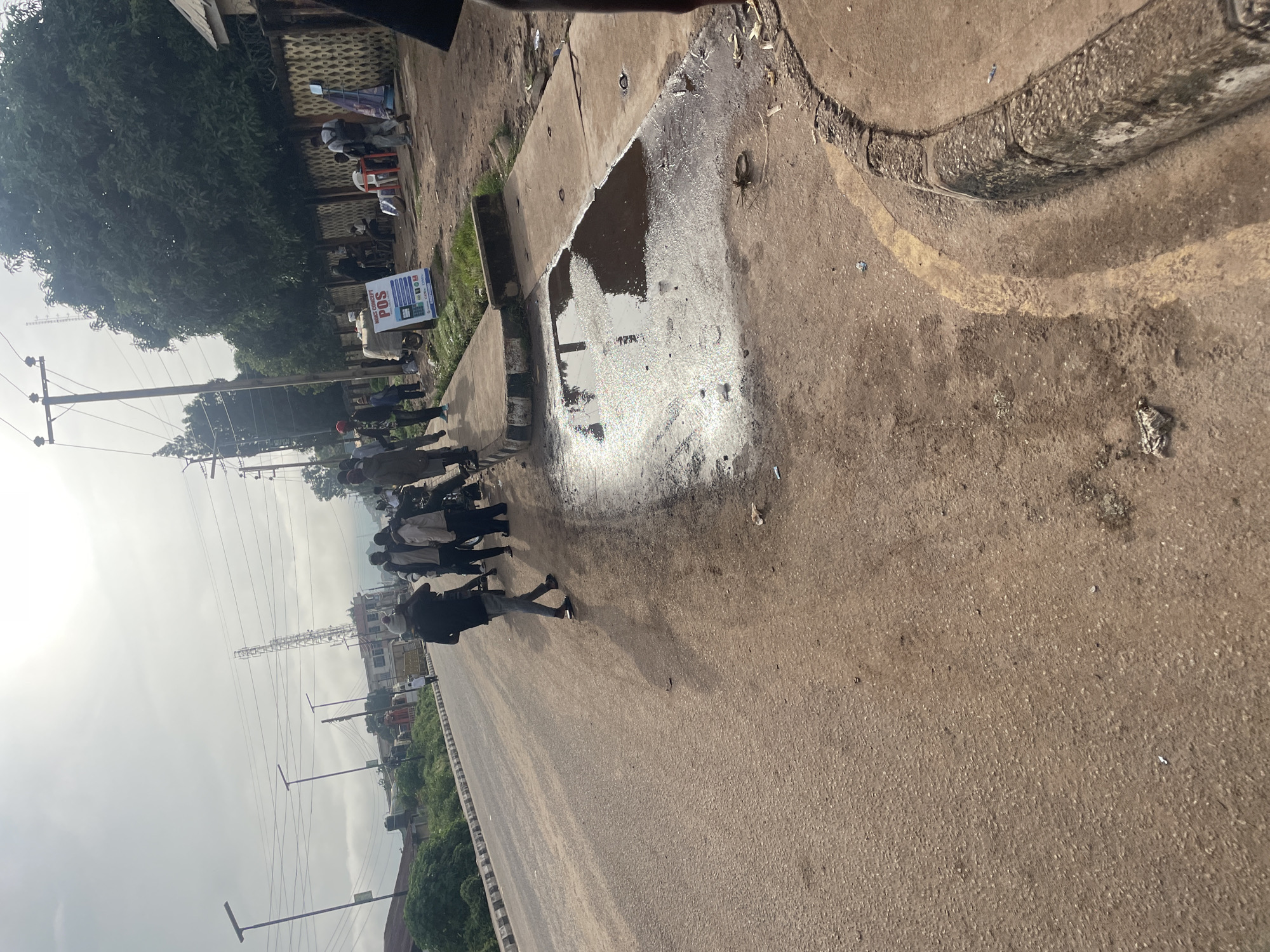 This image depicts a street scene in Jos, Nigeria. A group of individuals are gathered on the side of a wide, paved road. Several people are standing, some appearing to be in conversation or waiting. A large puddle of water is present on the paved area near the curb, reflecting the overcast sky and surrounding trees. A "POS" sign is visible on the right side of the frame, suggesting commercial activity. In the background, there are trees, buildings, streetlights, and a telecommunications tower, indicating an urban environment. The road is unpaved in some areas, with visible dirt and debris. The overall atmosphere appears to be a typical street scene in a Nigerian city.
