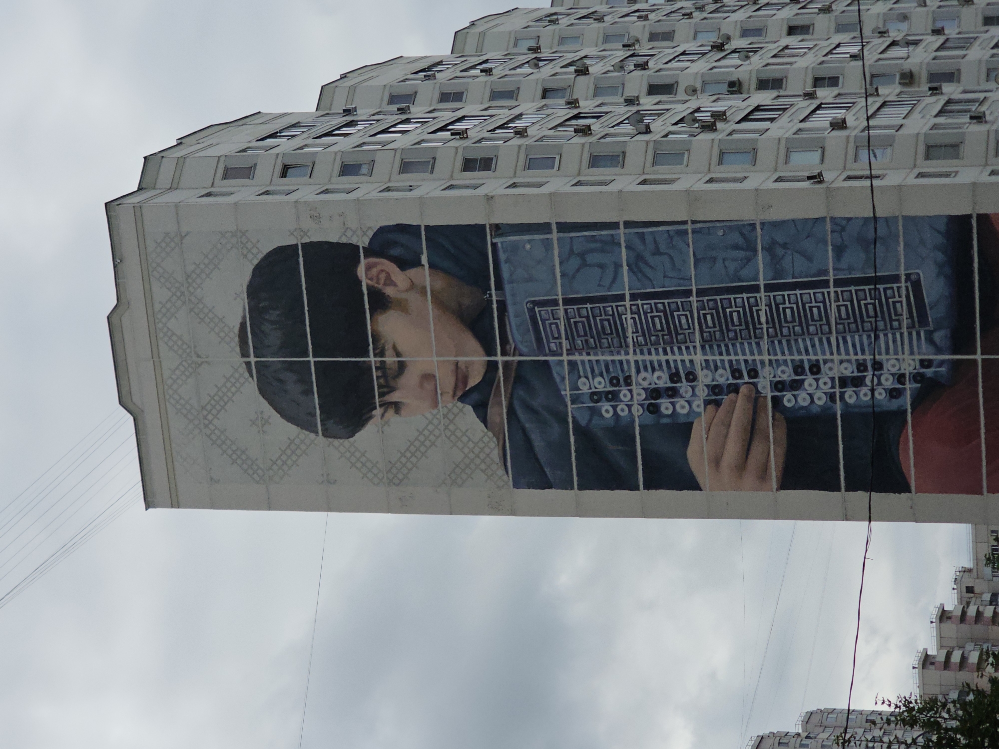 The image shows a mural painted on the side of a tall, multi-story apartment building. The mural depicts a young person with dark hair playing an accordion. The building itself is white with many windows and air conditioning units visible. The mural is divided into sections by vertical and horizontal lines, giving it a grid-like appearance. The sky is overcast and gray, with power lines strung across the scene. In the background, other apartment buildings are visible. The mural is located on a building in Odintsovo, Russia.