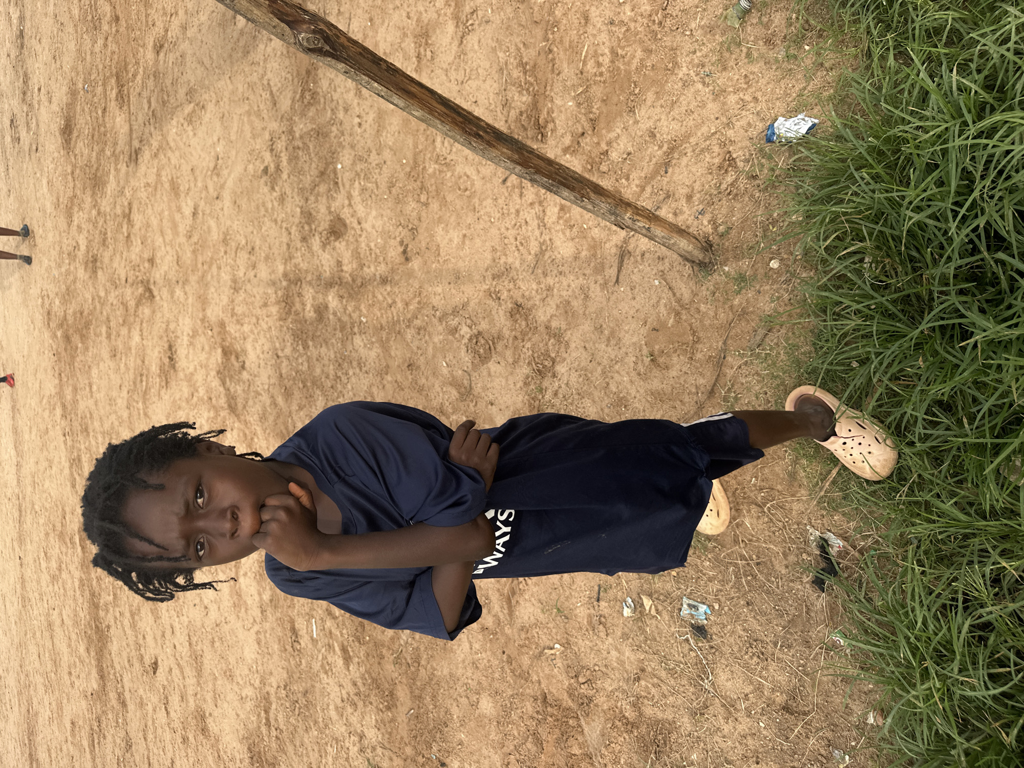 A young child with dark skin and braided hair stands in a dirt field with sparse green grass. The child is wearing a dark blue t-shirt with white lettering and dark shorts. They have their hand to their mouth and are looking directly at the camera with a serious expression. The child is wearing tan-colored slip-on shoes. A wooden pole stands to the right of the child. The ground is dry and sandy with some scattered litter. The location appears to be an outdoor public space, possibly a park or a sports field in Rukuba Road, Nigeria.