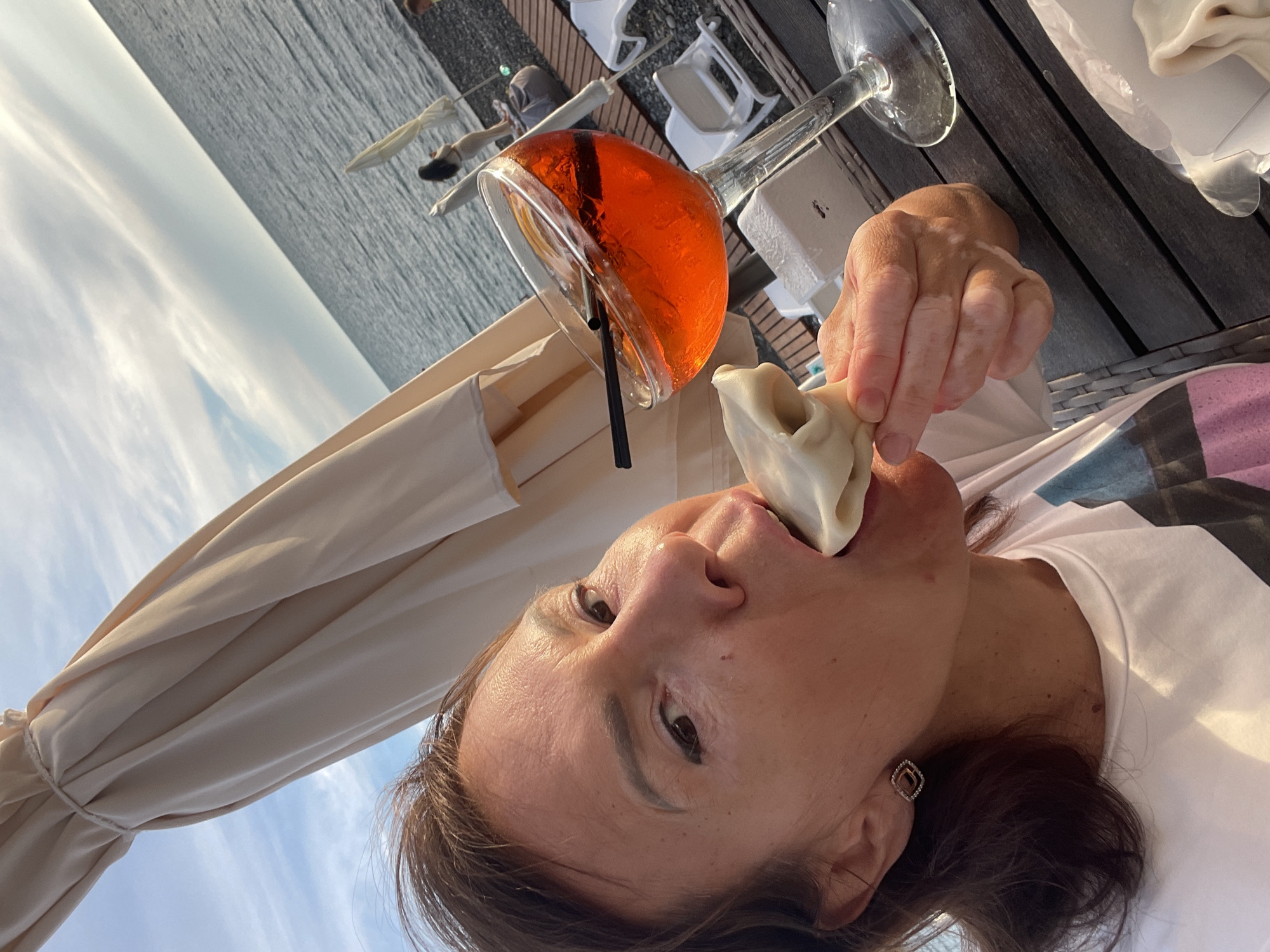 A woman is eating a dumpling while sitting at a wooden table outdoors at a beachside establishment. She is wearing a white t-shirt and a small, square earring with diamond accents. To her right is a large glass filled with an orange cocktail, possibly Aperol Spritz, with ice, an orange slice, and a black straw. In the background, the sea stretches to the horizon under a partly cloudy sky. Beach chairs and umbrellas are visible on the pebble beach. The scene suggests a leisure setting, likely on a warm day. The location is a beach club in Sochi, Russia, given the architectural style of the furniture and umbrellas, and the commonality of such establishments in the city's coastal areas.