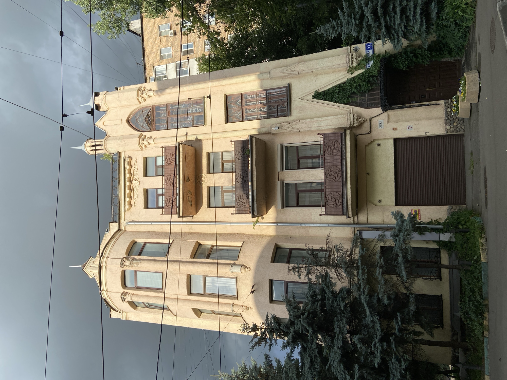 The image shows a multi-story building with an ornate facade, located on Baumanskaya Street in Moscow, Russia, as indicated by the street sign reading "БАУМАНСКАЯ 23". The building has a light-colored, possibly stuccoed exterior, with several balconies featuring decorative metal railings. The architecture includes a rounded corner with arched windows on the upper floors and a pointed, Gothic-inspired window on a tall, narrow section of the facade. The ground level features a roll-up garage door and a recessed entrance with a wooden door, framed by overgrown ivy. Two concrete planters with flowers are situated in front of the entrance. Evergreen trees, likely fir trees, flank the building on the left and right. The sky is dark and overcast, suggesting impending rain or a storm. Electrical wires are visible crisscrossing the sky in the foreground. A neighboring building, constructed of brick, is partially visible to the right. The overall scene depicts an urban street with distinctive architecture under dramatic weather conditions.