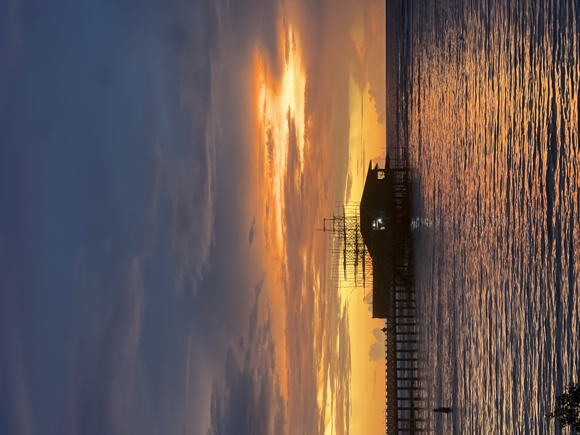 The image captures a sunset over the waters of General Luna, Philippines. The sky is ablaze with vibrant orange and yellow hues from the setting sun, interspersed with dark, brooding clouds. In the mid-ground, a stilted structure, possibly under construction due to the scaffolding, stands silhouetted against the colorful sky. A long wooden pier extends from the left side of the frame towards the structure, with a lone figure walking on it. In the foreground, the rippling water reflects the fiery colors of the sunset, creating a shimmering, golden surface. A person stands partially submerged in the water on the left side of the frame. The overall scene depicts a serene, yet dramatic, end to the day in this coastal Philippine city.