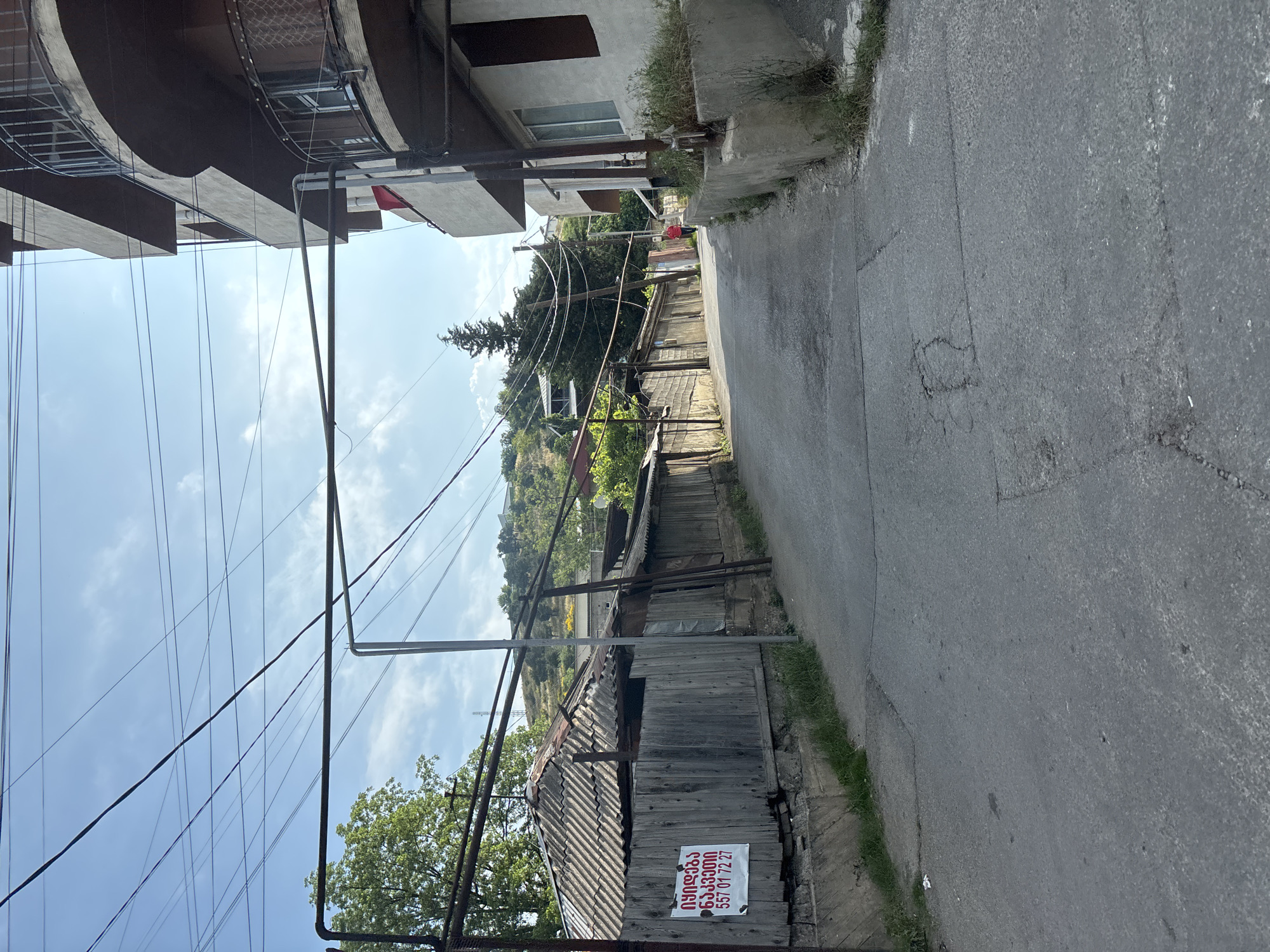The image depicts a narrow, asphalt road winding uphill through a residential area in Tbilisi, Georgia. The road is lined with a mix of older, weathered wooden structures with corrugated metal roofs and a more modern, multi-story building with balconies. Overhead, a complex network of electrical wires crisscrosses the sky. A sign with Georgian text and a phone number is affixed to the wooden fence of one of the older structures. In the distance, a person in a red jacket is walking uphill. The environment appears to be a typical urban street with elements of both older and newer architecture, suggesting a neighborhood with a long history.