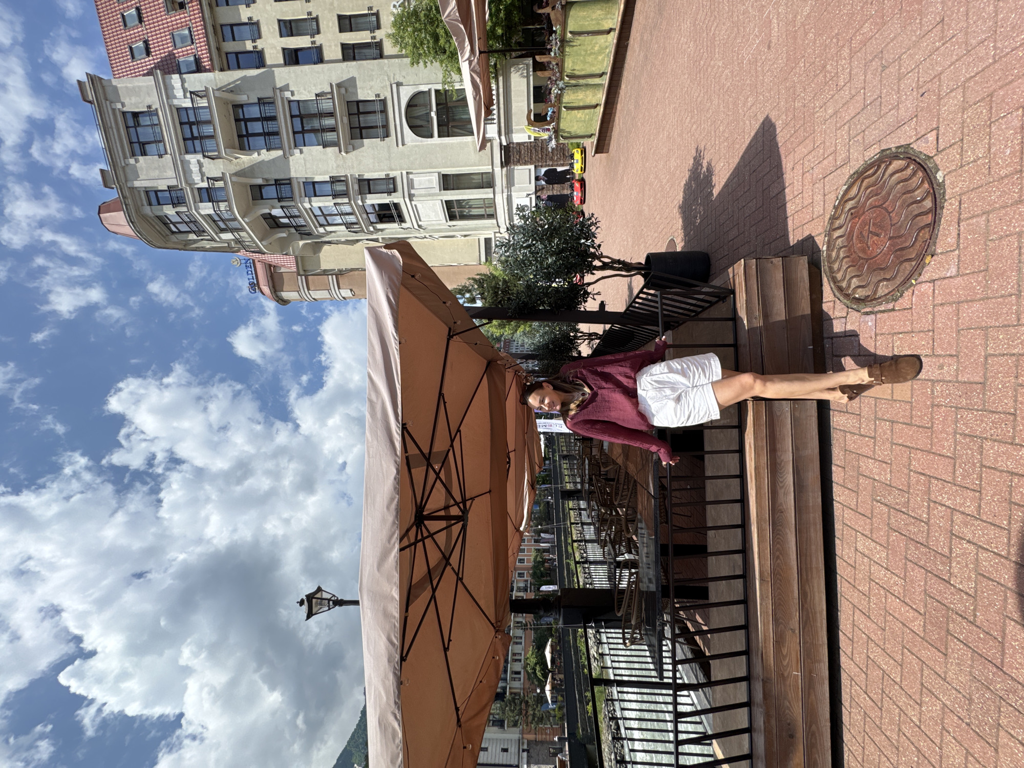 A young woman in a maroon button-down shirt and white shorts stands on a brick sidewalk in Estosadok, Russia. She is leaning on a railing near an outdoor seating area with umbrellas. Behind her, a river flows and a multi-story building with a red roof stands under a cloudy sky. In the distance, there are other buildings and trees. The scene suggests a pleasant, sunny day.