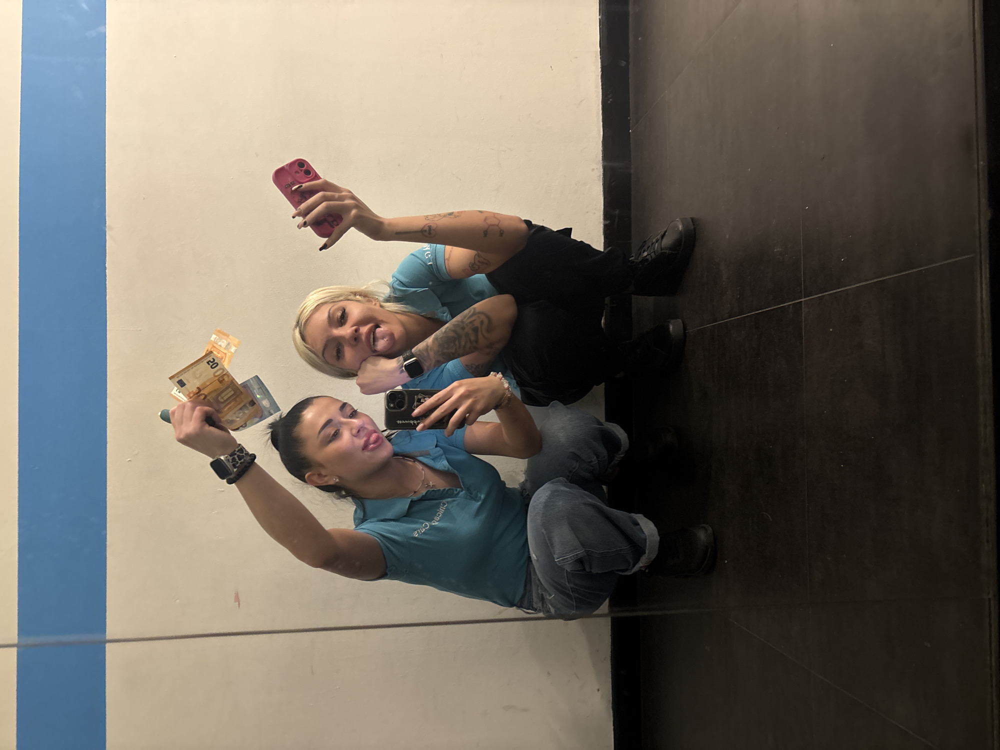 Two women are squatting, appearing to take a photo or video of themselves in a mirror. The woman on the left is holding Euro banknotes. Both women are wearing casual clothing. One is wearing a blue polo shirt and jeans, while the other is in a blue shirt and dark pants. The background includes a wall with a blue stripe and a dark-tiled floor. The photo was taken at a location in the city of Plovdiv, Bulgaria.