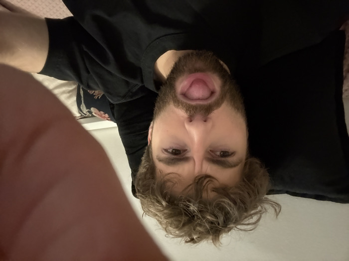 A bearded man wearing a black t-shirt looks directly at the camera, sticking his tongue out. He has curly blond hair. The photo appears to be taken from a close-up, upside-down perspective, possibly a selfie. A finger is visible in the bottom left corner. The man seems to be lying down, with a pillow behind his head.