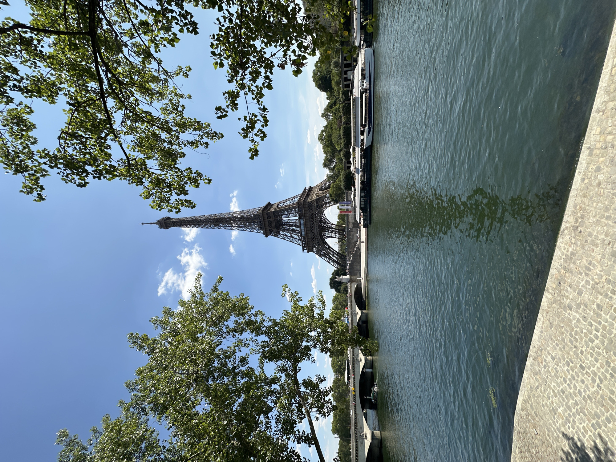 The image depicts a view of the Eiffel Tower in Paris, France, from across the Seine River. The Eiffel Tower is centrally located in the background, its iconic lattice structure clearly visible against a bright blue sky with scattered white clouds. In the foreground, the Seine River flows, with its surface rippling and reflecting the sky and surrounding elements. Along the riverbank, there is a paved walkway made of cobblestones. On the opposite bank of the river, a bridge spans the water, with traffic and pedestrians visible. Several boats are docked or cruising on the river, including larger tour boats. Lush green trees frame the scene from the left and right, their branches and leaves partially obscuring the view of the Eiffel Tower and the sky. The overall lighting suggests it is daytime, likely a sunny afternoon. There are no discernible individuals in the foreground, but there are some figures visible on the distant riverbank and the bridge.