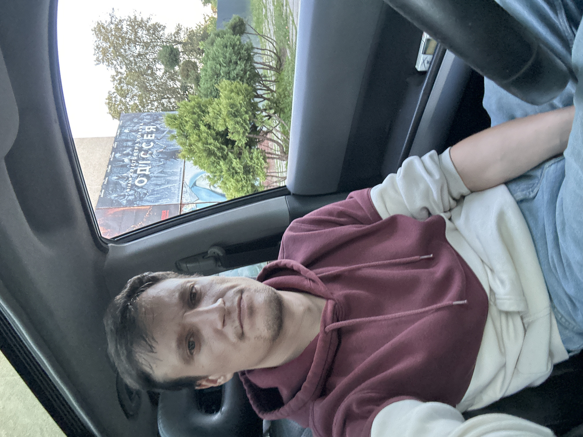 A man is sitting in the driver's seat of a car, taking a selfie. He is wearing a maroon and cream hooded sweatshirt and blue jeans. The car's interior is visible, including the steering wheel and dashboard. Through the car window, a large billboard for the movie "Odyssey" is visible, along with trees and a building. The billboard is located in Kamianets-Podilskyi, Ukraine.