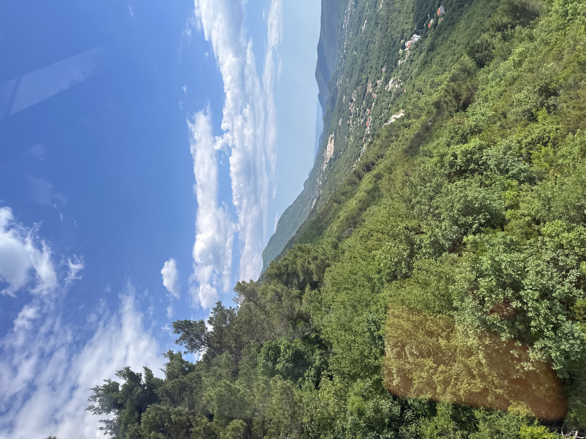 The image shows a landscape view, likely taken from an elevated position in or near Čavori, Montenegro. The sky occupies a significant portion of the frame and features a bright blue color with scattered white clouds. Below the sky, a mountain range is partially visible, with a portion of the Adriatic Sea appearing in the distance. The slope of a densely forested mountainside is the primary subject. The foliage is predominantly green, indicating lush vegetation. Scattered buildings and residences are visible on the mountainside. There is a visual obstruction on the left side of the sky.