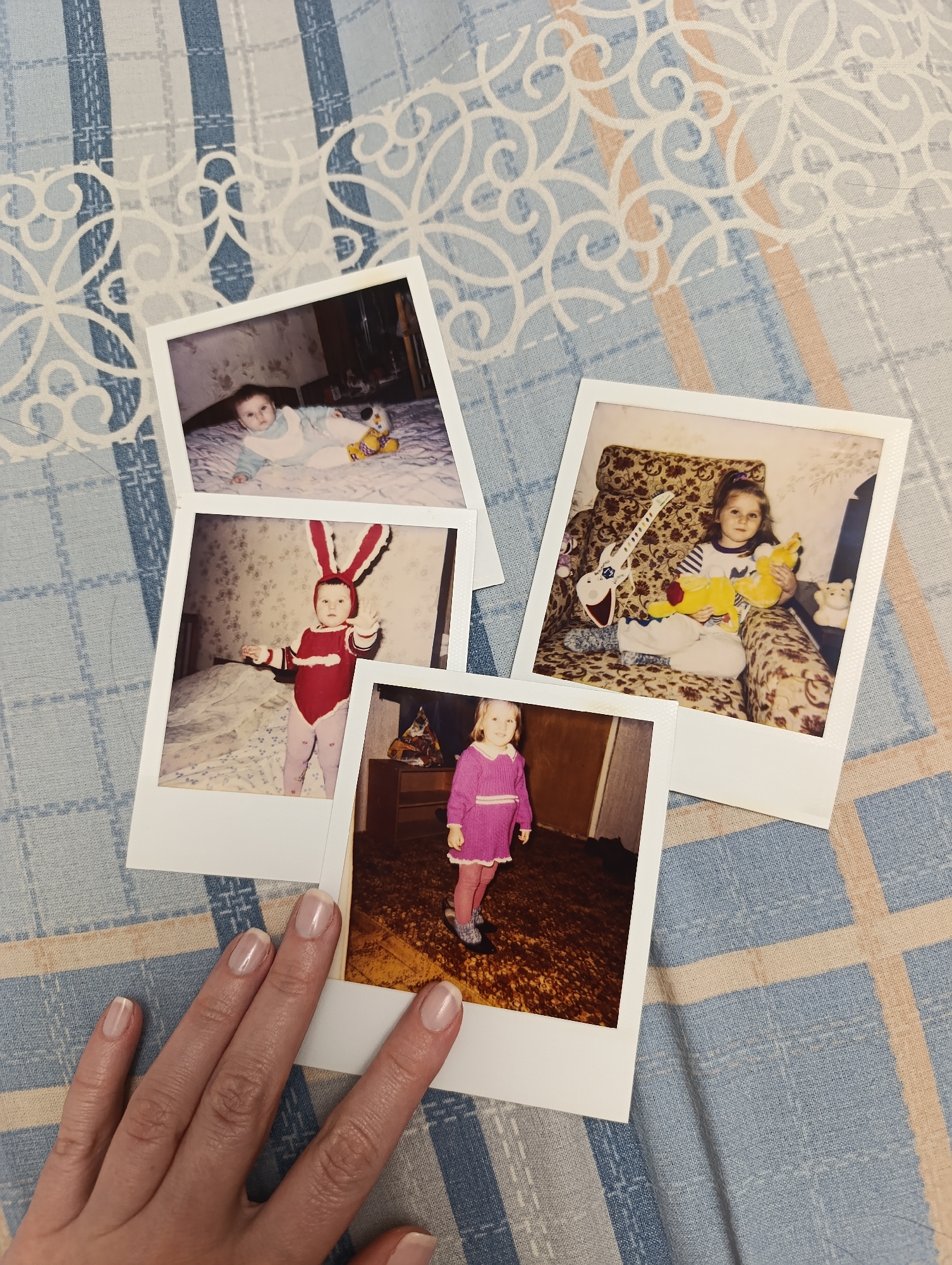 Four instant photos are arranged on a blue and tan patterned cloth surface, partially obscured by a left hand. The cloth features a grid pattern with floral accents.
The instant photos depict a young child at different ages. From top left, clockwise:
1. A baby on a bed with a toy.
2. A young girl in a patterned armchair holding a stuffed animal and guitar.
3. A young girl in a pink outfit standing in a room with a wooden dresser.
4. A young child in a red and white bunny costume standing in a bedroom.
A left hand with pale skin and manicured nails rests on the bottom of the photo stack.