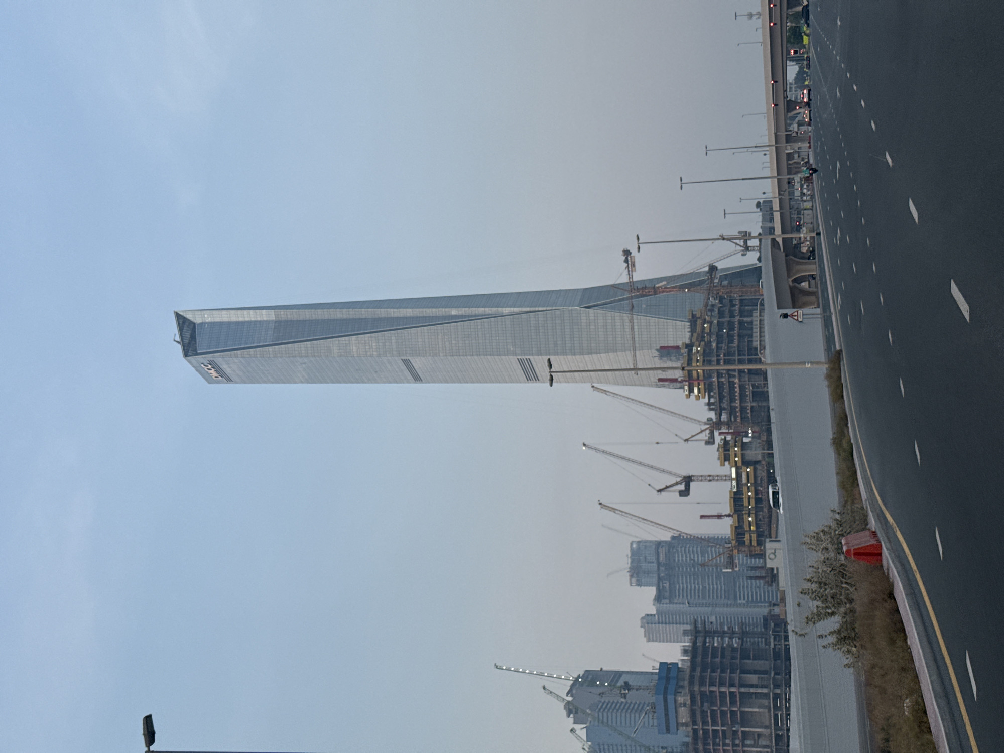 This is an eye-level, medium shot of a modern city street scene in Dubai, United Arab Emirates, under a pale, overcast sky. The dominant feature is a very tall, modern skyscraper with a unique, angled design, likely under construction, with the letters "DMCC" visible at its apex. In the foreground, a dark asphalt road runs from the bottom left towards the right side of the frame, with white dashed lines marking the lanes. A yellow line is visible on the left edge of the road. To the left of the road, a gray concrete barrier runs along the edge, with a small patch of dry, brown vegetation and a red traffic cone on the roadside.
Behind the barrier, several other buildings are under construction, identifiable by scaffolding, cranes, and exposed structural elements. Multiple large construction cranes are visible throughout the midground. Further back, other completed or near-completed modern buildings can be seen, contributing to the urban skyline. Several streetlights line the road and the elevated highway overpass visible in the distance. A white van is visible on the road behind the barrier, and several cars are present on the elevated highway. The overall impression is one of urban development and ongoing construction in a large city.