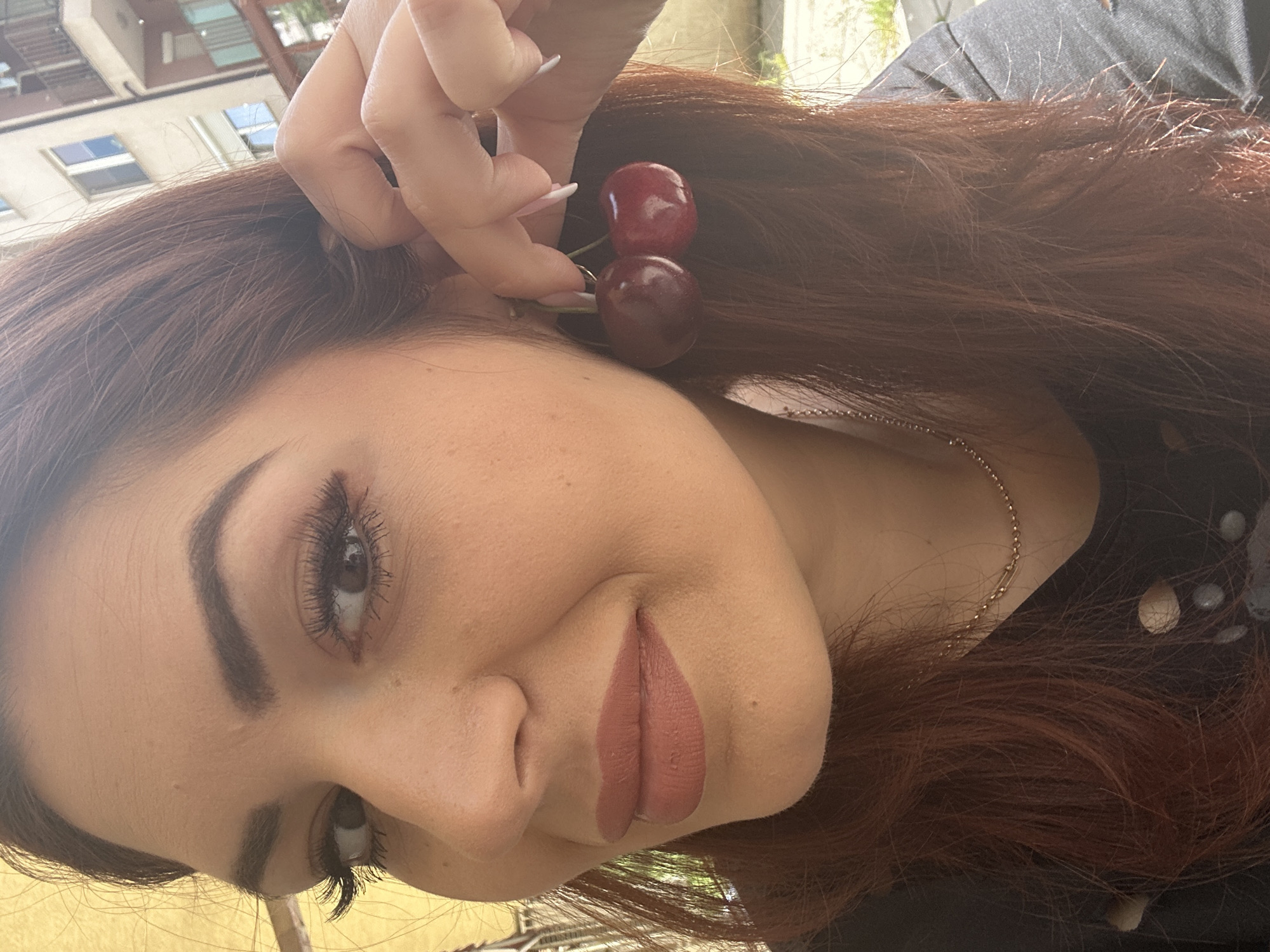 A young woman is holding two cherries near her ear. She has dark auburn hair and is wearing a thin silver chain necklace and a dark shirt with white polka dots. She is wearing makeup, including lipstick and mascara. The background includes urban architecture, suggesting an outdoor location. Considering the given information, it is likely that this photo was taken in the city of Sofia, Bulgaria.