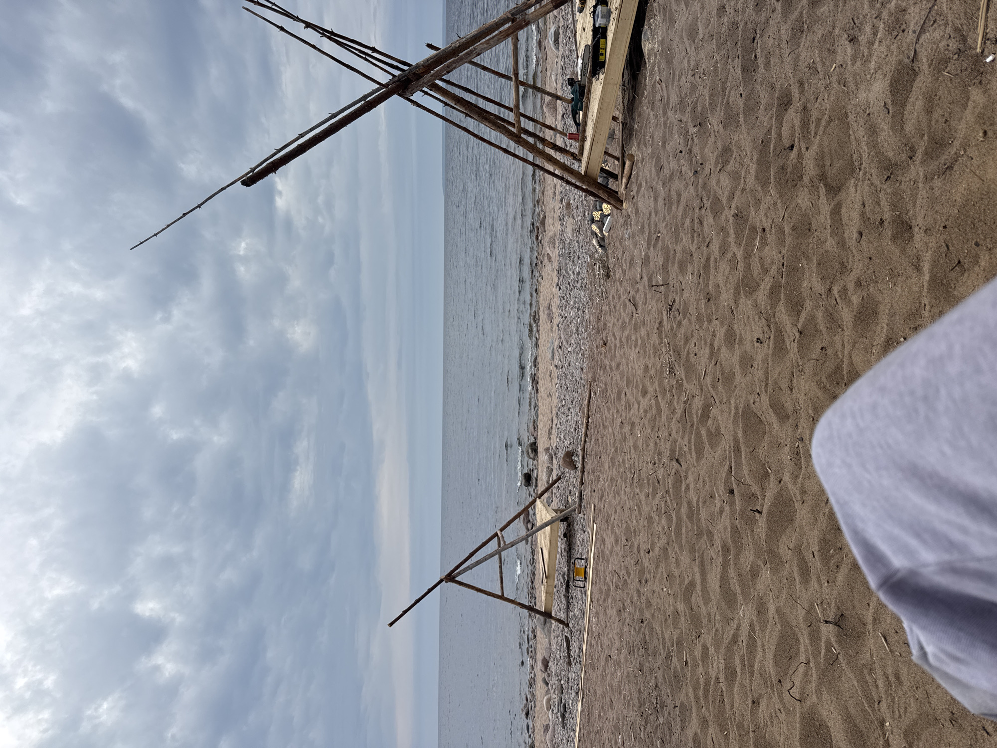 The image depicts a beach scene in Baltiiskoe, Russia. The view is angled, showing the beach in the foreground and the sea and sky in the background. Two wooden structures are visible on the beach. The structures appear to be A-frame or tripod-like, constructed of wooden poles. One structure is taller than the other. The beach consists of sand and small rocks. The sky is overcast with clouds. The sea is calm. Part of a white or gray object, possibly clothing, is visible in the lower right corner of the image. There are no people visible in the main scene.