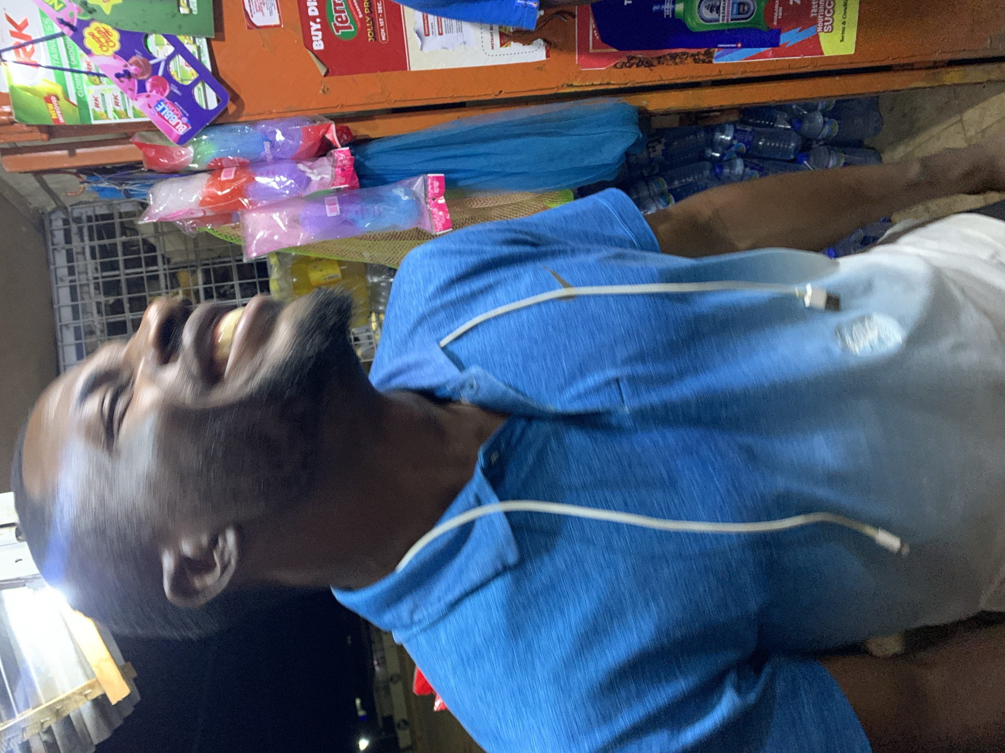 A man with dark skin, a beard, and a bald head is shown smiling broadly in a vertically rotated image. He is wearing a blue polo shirt with white drawstrings. Behind him, a partially visible stall displays various items including candy, bottles of water, and other packaged goods. A sign with text like "BUY. DIS" and "Terr" is visible at the top of the frame. It is difficult to provide more specifics about the location beyond that it appears to be a stall with a range of products, potentially in a bustling urban area.