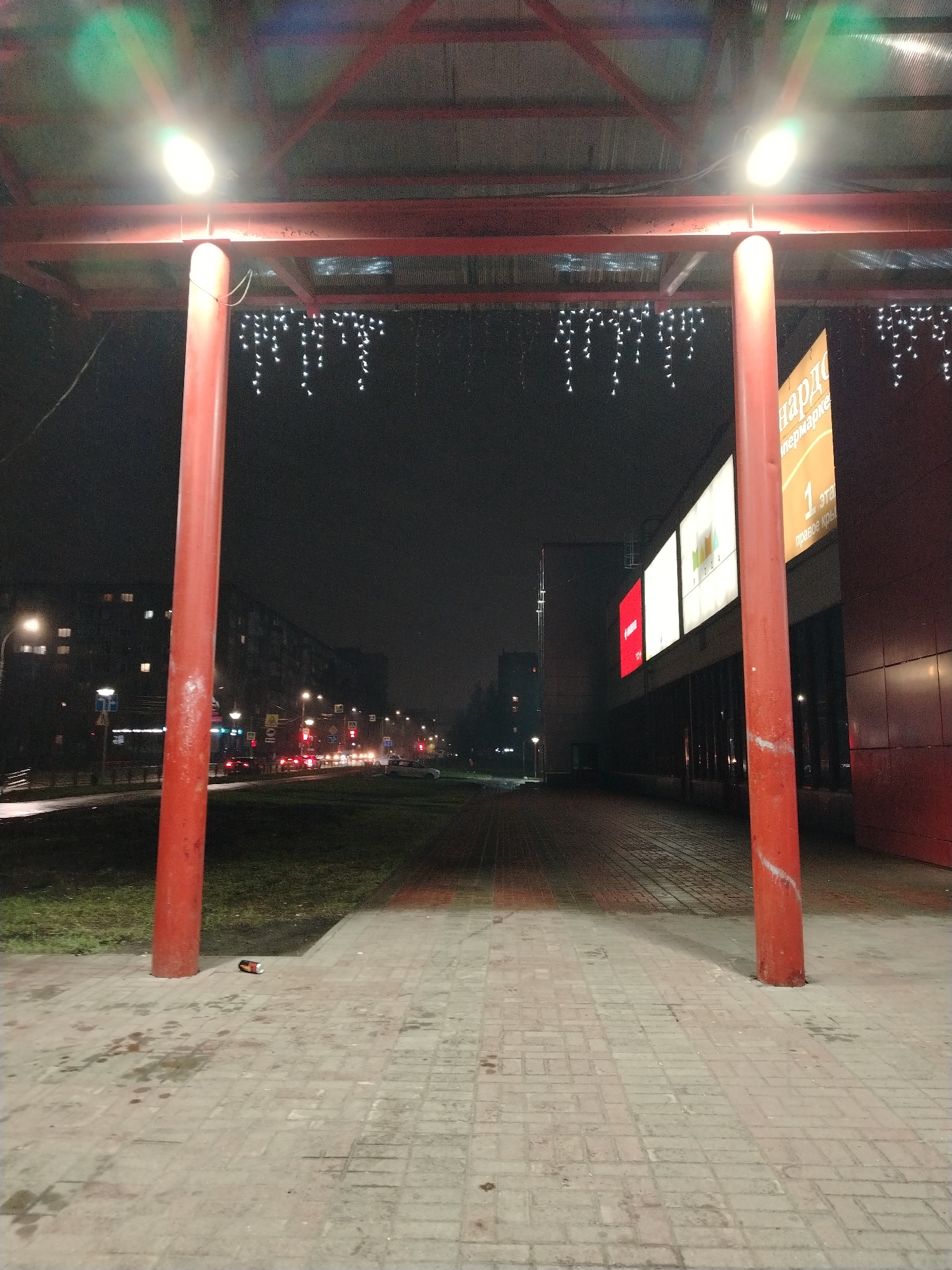 The photograph shows an outdoor scene at night in Izhevsk, Russia. The viewpoint is under a red-painted metal structure with two supporting cylindrical columns. Two illuminated lights are attached to the overhead structure. Under the structure are hanging decorative lights that resemble icicles. The ground is paved with light-colored bricks. There is a discarded can on the ground near the left column.
Beyond the structure, there is a grassy area bordering a road. Buildings line both sides of the road, with illuminated windows visible in the buildings on the left. Traffic, including car headlights and taillights, can be seen on the road.
To the right, attached to one of the buildings, are illuminated advertising signs. The nearest large sign has a predominantly orange background with Russian text. Adjacent to it are three illuminated billboards with white backgrounds. The building itself has a dark red facade with minimal illumination.