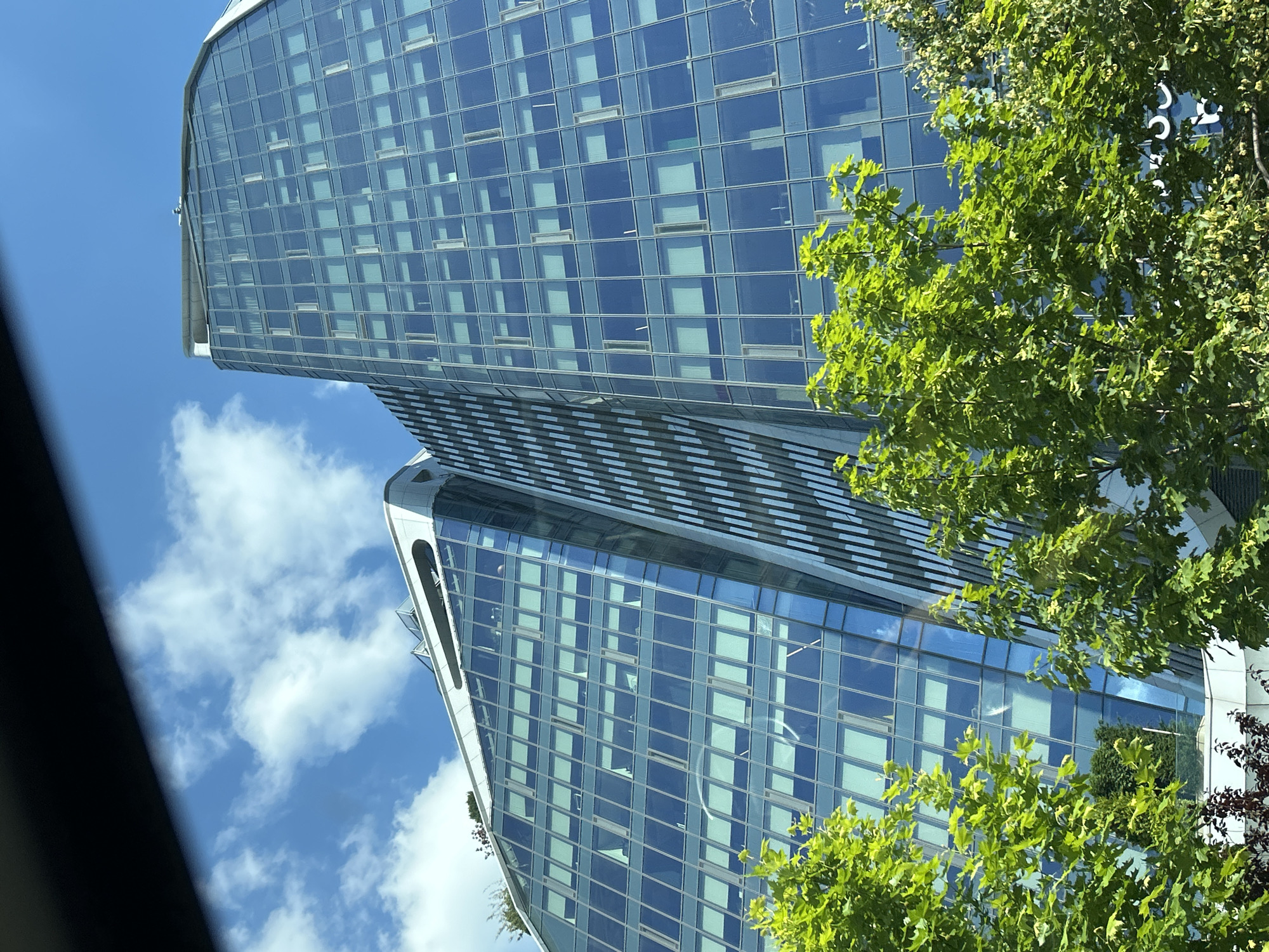 The image shows a modern, glass-fronted skyscraper against a clear blue sky with scattered white clouds. The building's facade is composed of numerous rectangular windows, some reflecting the sky and others appearing to be illuminated from within. The skyscraper has a unique architectural design, with curved sections and a sleek, contemporary appearance. In the foreground, lush green trees with dense foliage partially obscure the building, their leaves catching the sunlight. One of the trees has some dark red leaves mixed in.
The scene appears to be set during daytime, likely midday, given the bright sunlight and clear sky. The presence of the trees and the bright weather suggest an outdoor setting in an urban environment, possibly a business district or a park area adjacent to a city center. The image is captured from a low angle, looking upwards towards the towering structure and the sky. There is no discernible activity or event taking place, and no people are visible in the frame. The overall impression is one of architectural grandeur and a harmonious blend of nature and urban development. No text is visible in the image.