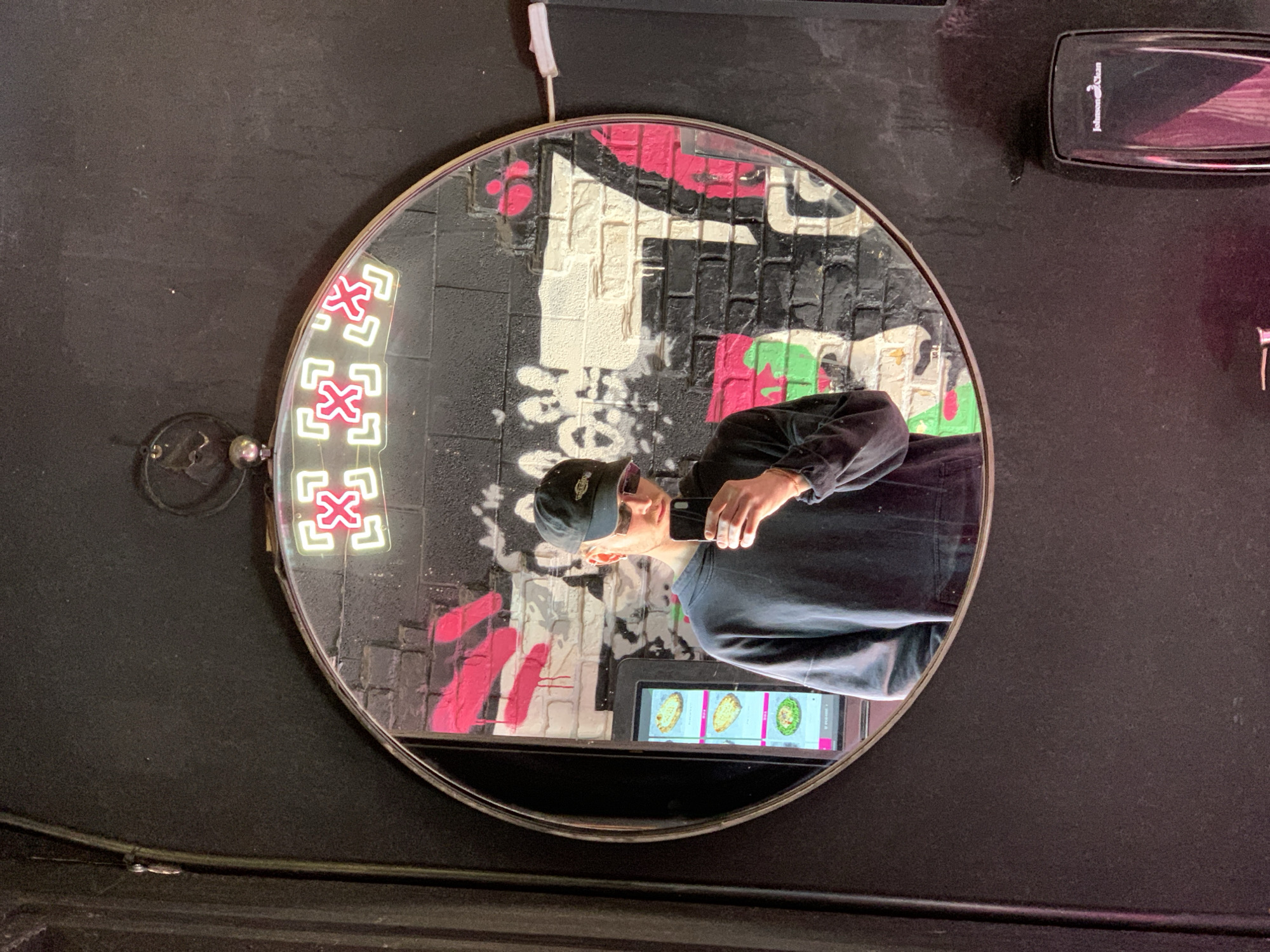 A man in a black baseball cap and black clothing is reflected in a round mirror mounted on a dark wall. The mirror reflects the man holding a smartphone, facing him. Behind the man in the reflection is a graffiti-covered wall with red, green, white, and black paint, as well as a neon sign composed of four stacked square symbols. The smartphone screen displays images of food items. Above the mirror, there is a white cable and a black rectangular object. The scene is reminiscent of a restaurant or food service setting. While the prompt mentions the location in Tel Aviv, Israel, this cannot be forensically validated based on the image alone.