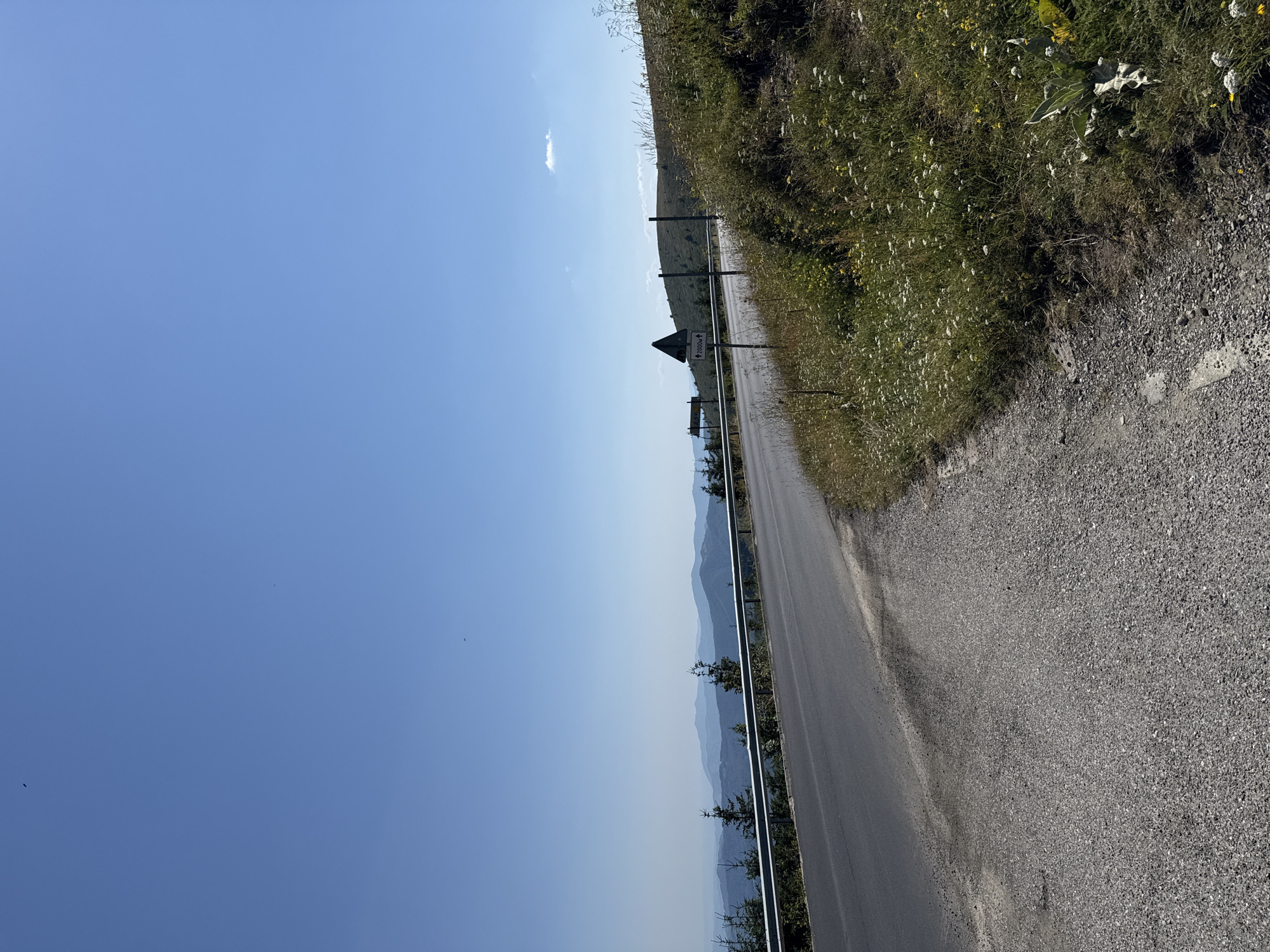 The image depicts a scenic view of a winding asphalt road in the mountainous region near Beli Osam, Bulgaria. The road is flanked by a metal guardrail on the left and a grassy embankment with wildflowers on the right. In the background, a vast expanse of blue mountains stretches towards the horizon under a clear, bright blue sky with a few wisps of clouds. The foreground shows the road surface with scattered gravel. A triangular road sign with an arrow and the text "2000M ↑" is visible near the roadside, indicating a distance of 2000 meters. The environment appears to be a natural, rural setting, likely a mountain pass or scenic route.