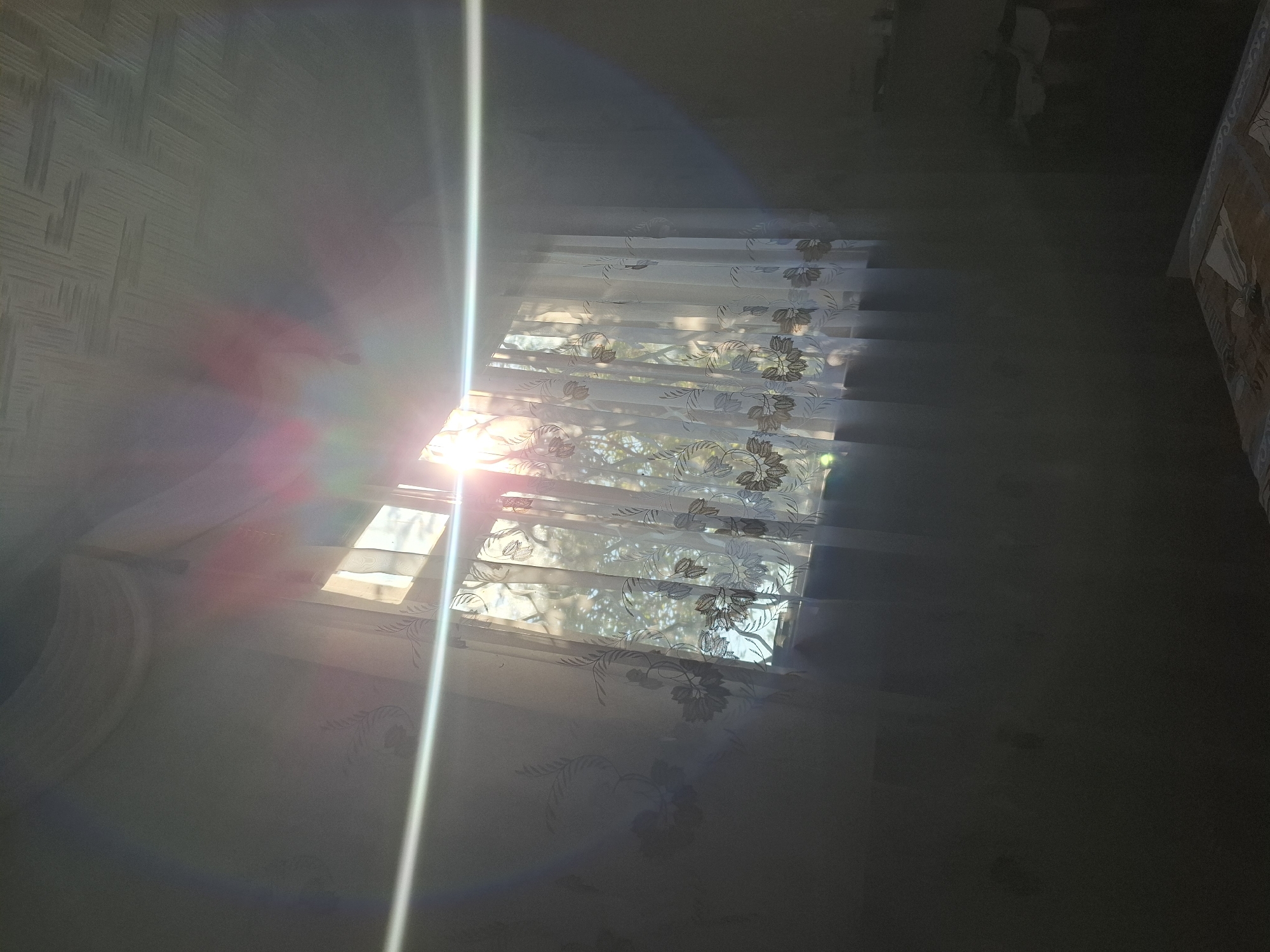 The image depicts an interior scene, likely a bedroom, with sunlight streaming through a window covered by sheer curtains. The curtains have a floral pattern. The sunlight creates lens flare effects, visible as streaks and circles of light across the image. The walls appear to be painted a light color, with some subtle floral wallpaper visible on the left side. The ceiling is textured. The lower portion of the image is darker, showing what appears to be a bed with patterned bedding. The overall impression is one of a domestic interior bathed in natural light. The location in Atkarsk, Russia is not visually discernible from the image itself.