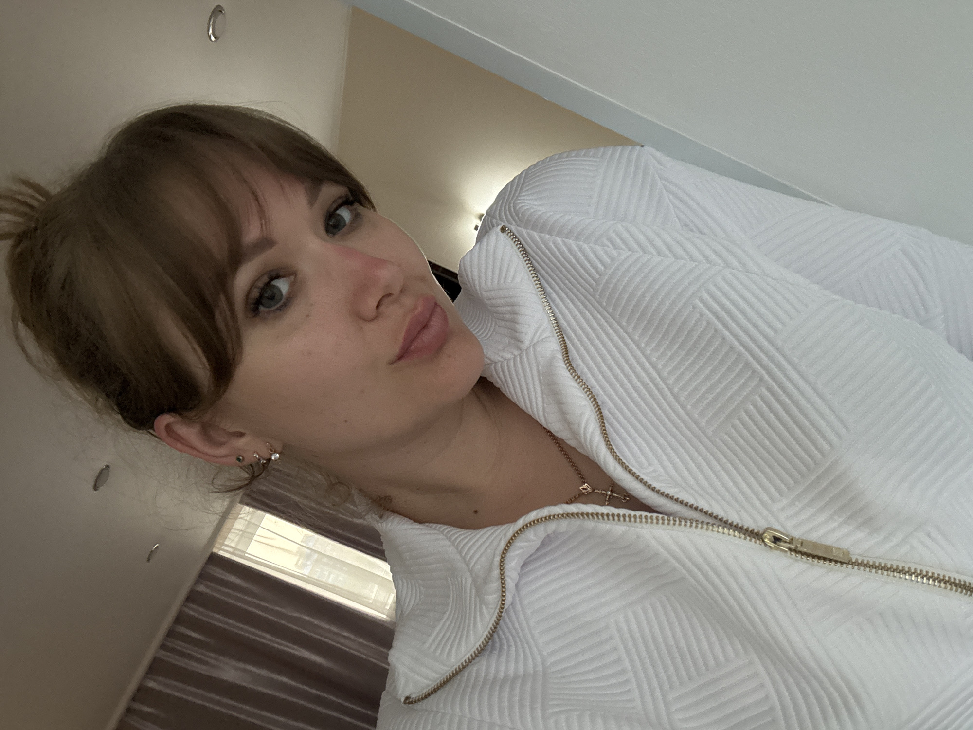 A woman with brown hair pulled into a bun and bangs is posing for a selfie. She is wearing a white textured jacket with a gold zipper. The background is blurry, but it appears to be an indoor setting with a window and light-colored walls. There is no definitive evidence to suggest the location is Yevpatoriya, Ukraine.