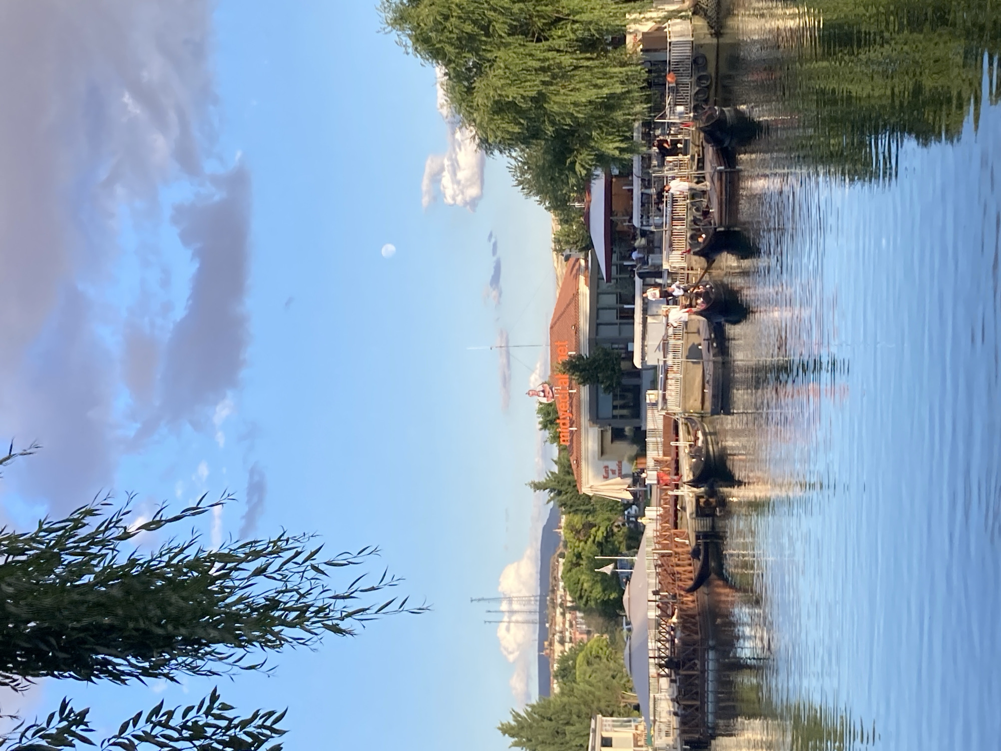 The image depicts a waterfront scene in Avanos İlçe Merkezi, Turkey. In the foreground, a tranquil blue body of water reflects the sky and surrounding environment. Several dark-colored gondolas are moored near a wooden pier and boardwalk. People can be seen standing on the boardwalk and near the water's edge, with some appearing to be waiting for or disembarking from the gondolas.
Behind the boardwalk, a row of buildings lines the waterfront. One prominent building features a large orange sign with Turkish text, possibly indicating a restaurant or establishment. Other structures include buildings with pitched roofs, awnings, and outdoor seating areas. Tall green trees, including a large willow tree with drooping branches, frame the scene on the right side. In the distance, hills are visible, topped with structures and communication towers, suggesting an urban or semi-urban landscape. The sky is a clear blue, with scattered white and gray clouds, and a small, pale moon is visible in the upper portion of the frame. The overall impression is a calm, daytime scene with leisure activities taking place by the water.