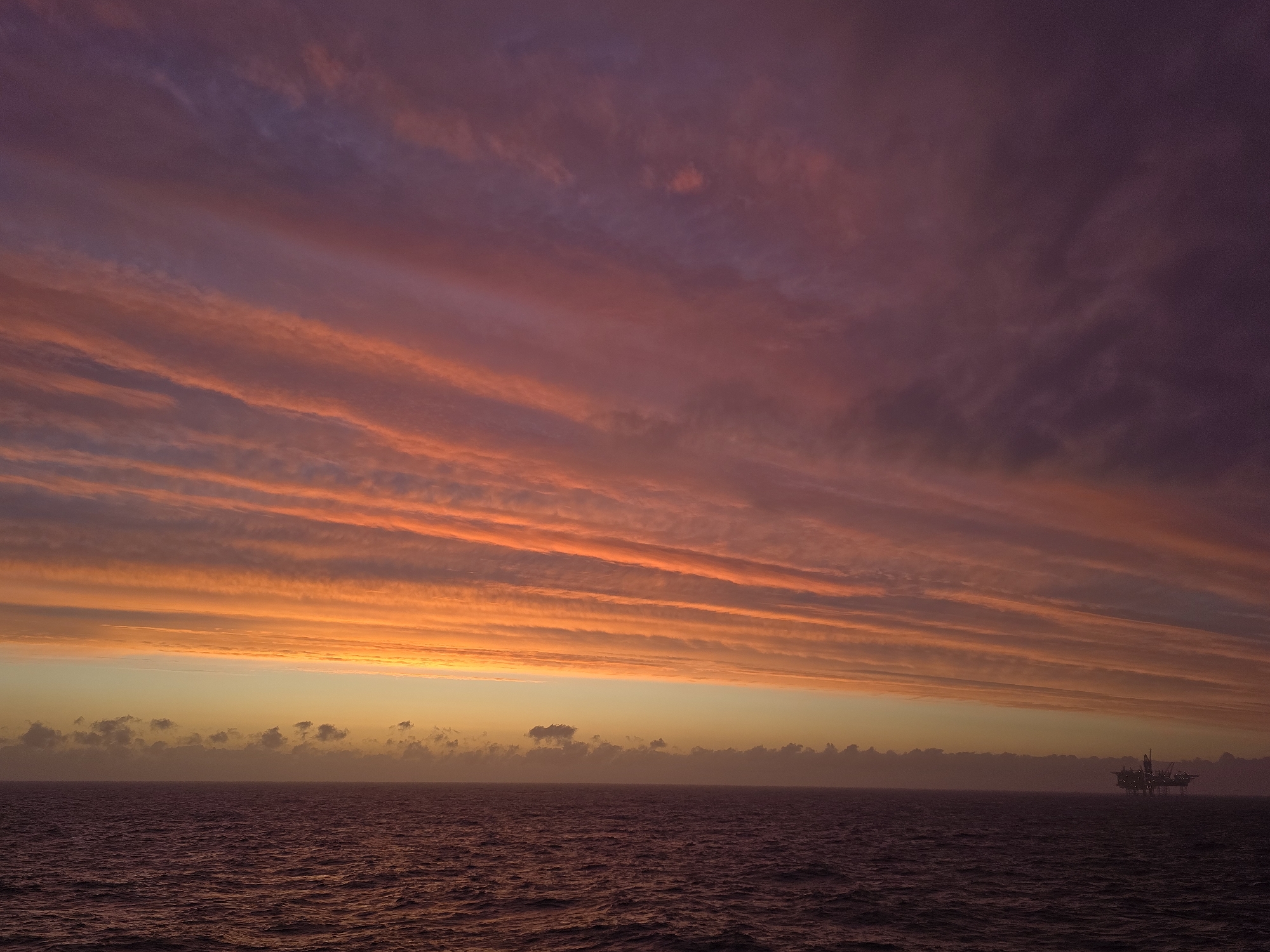 The image depicts a seascape under a vibrant, multi-hued sunset. The lower portion of the image is dominated by a dark, rippling body of water, likely the ocean. The horizon line is somewhat obscured by a low-lying bank of clouds, tinged with the colors of the setting sun. Above this, the sky transitions from a bright orange near the horizon to pink and purple hues higher up. The clouds are streaked horizontally across the sky, creating a layered effect. In the distance, on the right side of the image, there is a structure that appears to be an oil platform. The scene is set in Unknown, Unknown.