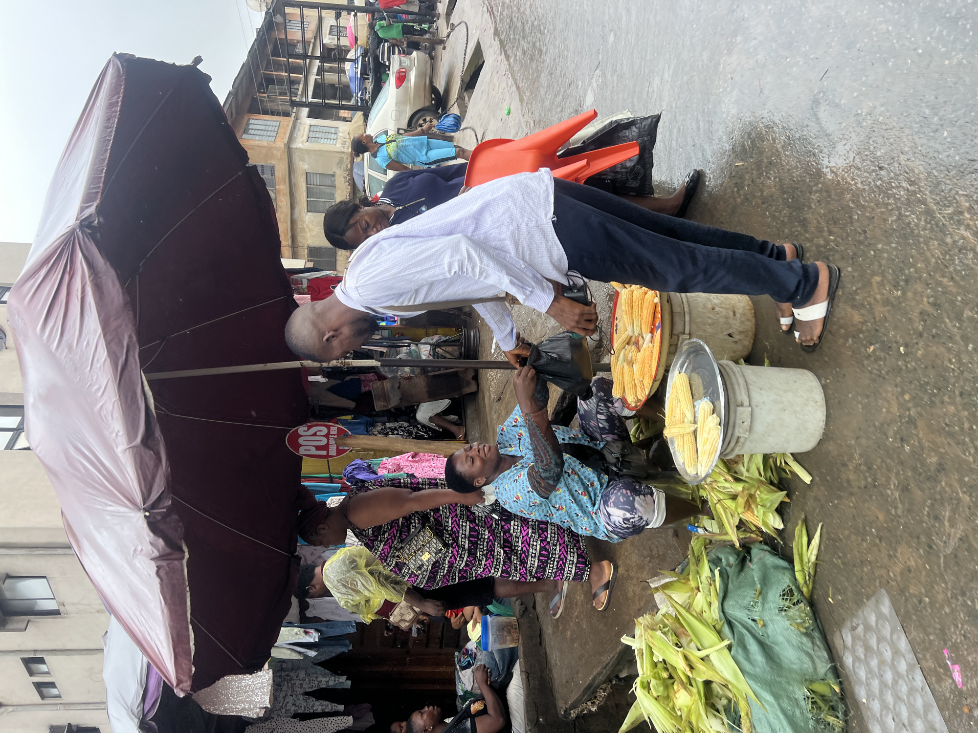The image depicts a street market scene in Onitsha, Nigeria. Under a large, worn, burgundy umbrella, a woman is selling corn. She is crouched down, wearing a blue patterned shirt and dark leggings, and is interacting with a man who is standing and reaching out to take a plastic bag of corn. The man is wearing a white long-sleeved shirt and dark trousers. In the foreground, a large pile of husked corn is visible on a green tarp. Behind the woman, a metal tray of corn is placed on a white bucket. Other market stalls and people are visible in the background, as well as buildings lining the street. The ground is wet, suggesting it may have recently rained or is raining. The overall atmosphere is one of active commerce in an outdoor urban setting.