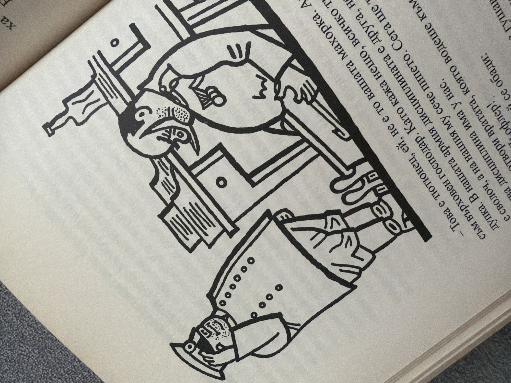 The image is a close-up, overhead shot of an open book with a line drawing illustration. The illustration depicts two cartoonish figures and a table with a bottle. One figure, dressed in a military-style uniform with a hat, stands at the left, saluting. The second figure, wearing a coat and a round object pinned to their chest, is seated at the table on the right, with their legs extended. The table has a bottle and what appears to be papers or books on it. The text below the illustration is in Cyrillic script. There are no identifiable landmarks of Pazardzhik, Bulgaria in the image.