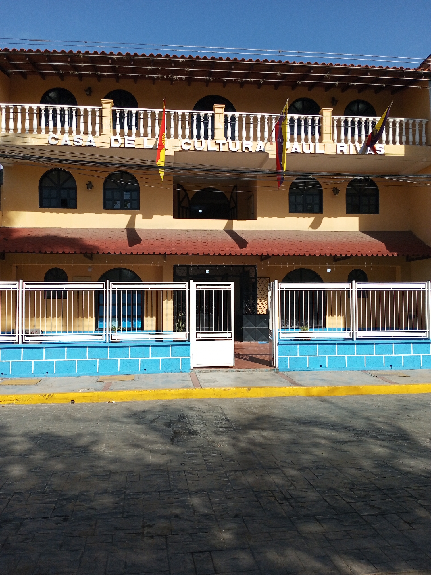 The image shows the "Casa de la Cultura Paul Rivas," a cultural center in Palo Negro, Venezuela. The building is two stories high and painted a yellow-orange color. There is a white-railed balcony on the second floor, with the name of the center displayed in white letters. Two flags of Venezuela are hung. The building's roof is red. A white metal fence surrounds the front of the building, with a blue base. A yellow curb runs along the front of the building, separating it from a paved area. The day is sunny, as evident from the shadows on the ground.