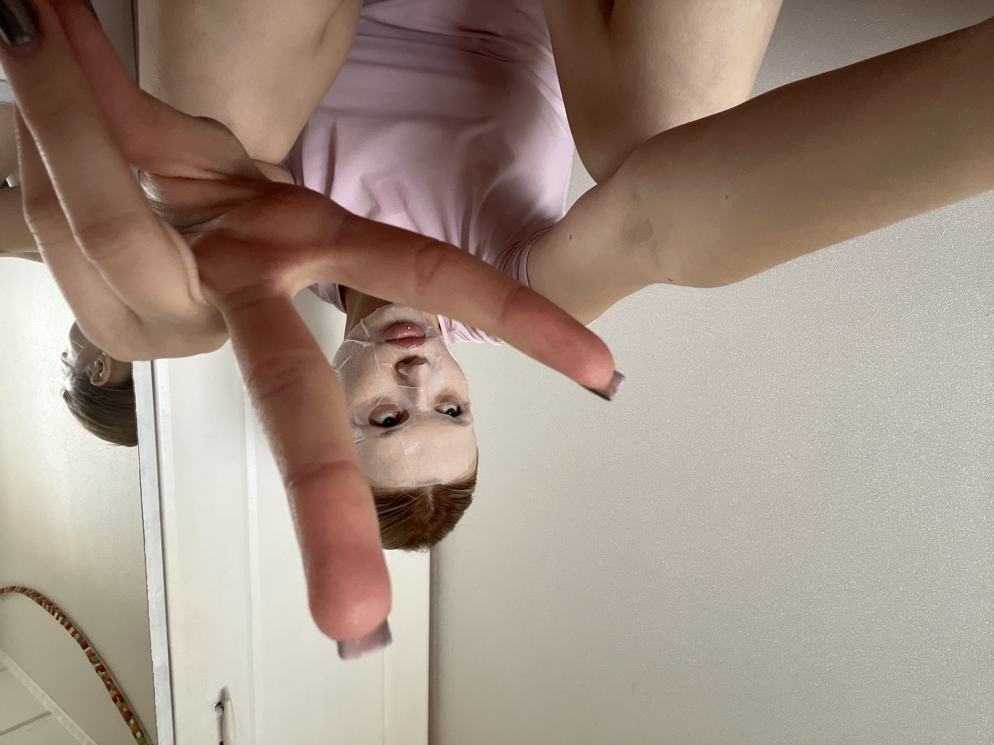 The image captures a unique, low-angle perspective of a person engaging in a self-care routine indoors. The main subject is a person, likely female, with reddish-brown hair, seen from below as if taking a selfie. Her face is covered with a white sheet mask, with her eyes open and looking directly towards the camera, and her lips slightly parted. She is wearing a light pink or lavender short-sleeved top. Her hands are prominently featured in the foreground, extended towards the camera, with some fingers sporting dark-colored nail polish. Her arms and upper torso are visible, with legs extending upwards out of frame.
The setting appears to be an indoor room, possibly a bathroom or bedroom. On the left side of the frame, a reflective surface, likely a mirror, shows a reflection of the person's head and hair. Also visible in the reflection is a partial view of a colorful, striped hula hoop or similar circular object, and a white door or cabinet. The right side of the background consists of a plain, light-colored (possibly white or very light grey) wall with a subtle textured finish. The image is taken from a perspective looking upwards at the person, who seems to be positioned above the camera, possibly lying down or leaning back, creating an unusual, inverted composition. There is no visible text in the image. The time of day and external weather are not discernible. No specific visual cues indicate the location as Chaikovsky, Russia.